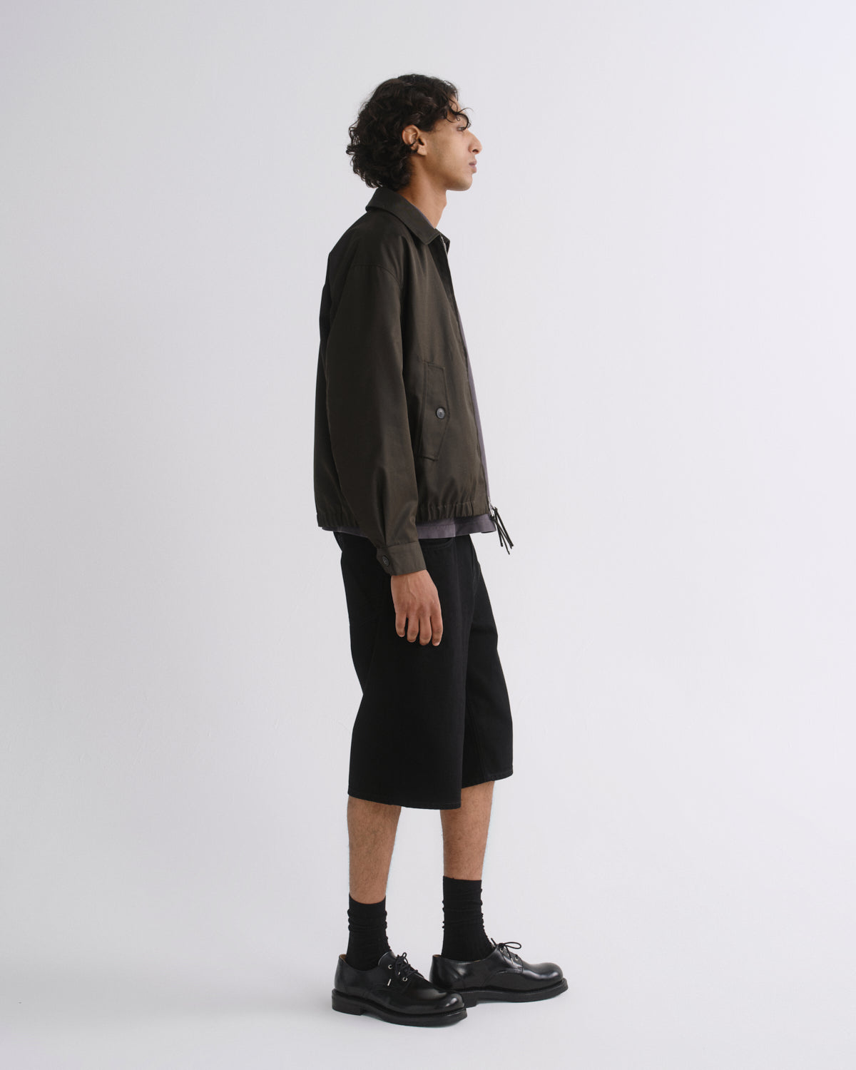 A man in side profile wearing a dark olive-brown utility jacket, black shorts, black socks, and black leather shoes, standing against a light gray studio background.
