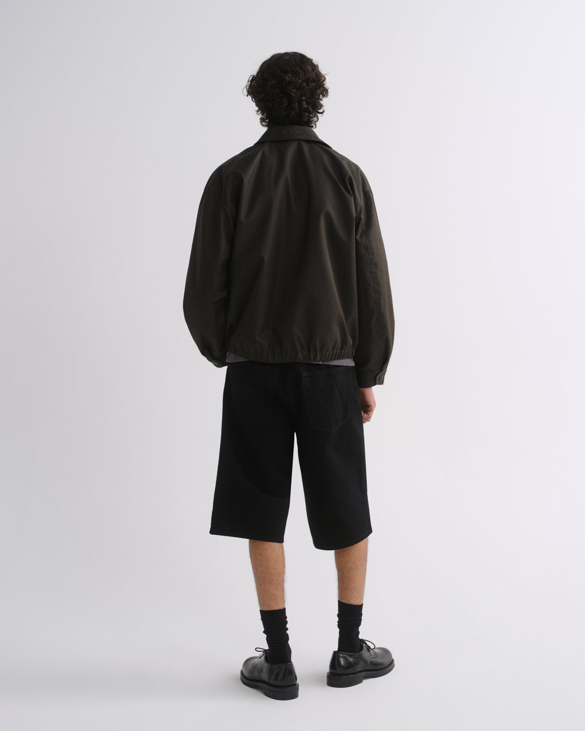 A model shown from the back wearing a dark olive-brown jacket, black shorts, black socks, and black shoes against a light gray studio background.