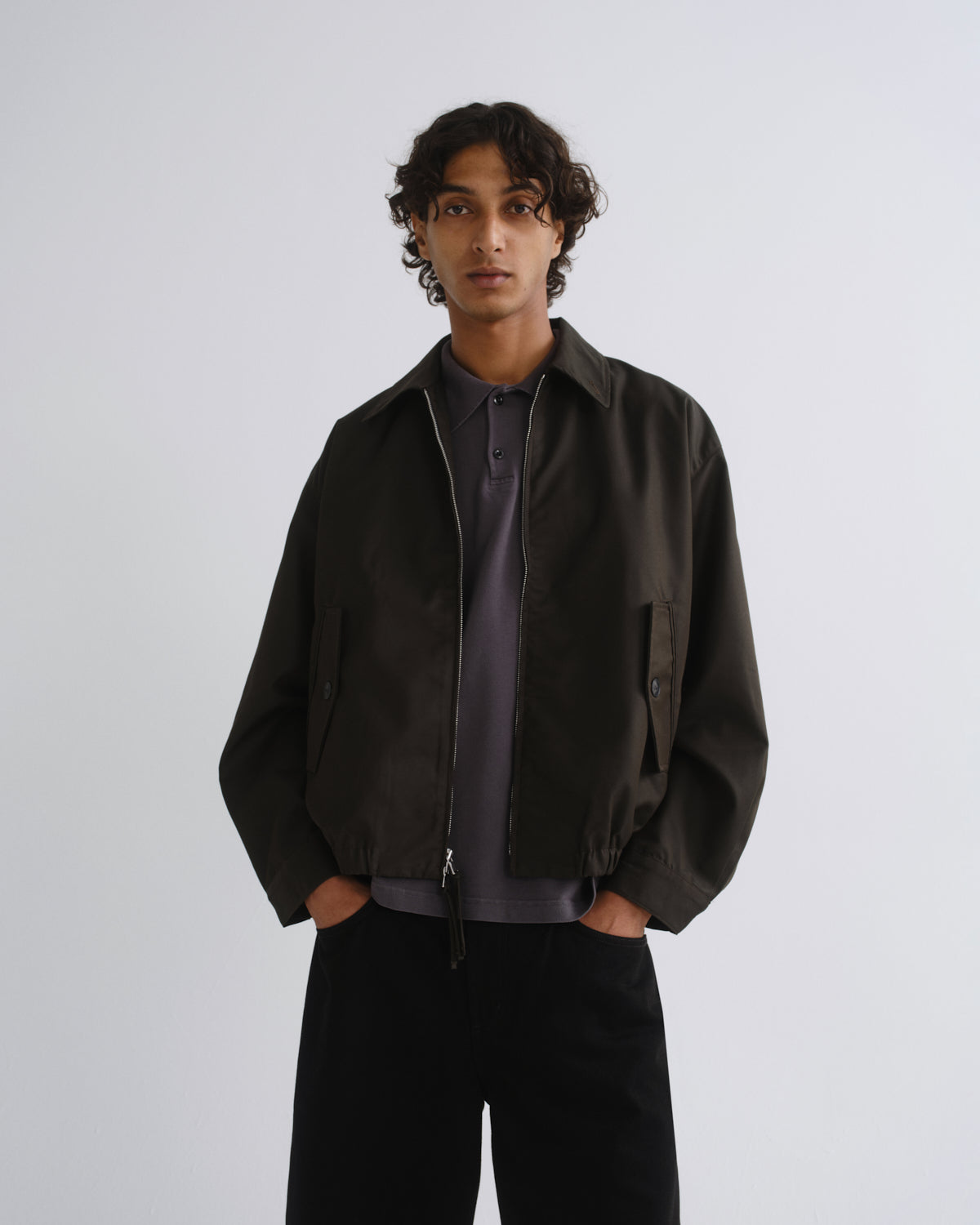 A man wearing a dark brown zip-up jacket over a dark gray polo shirt and black pants, standing against a light gray studio background.