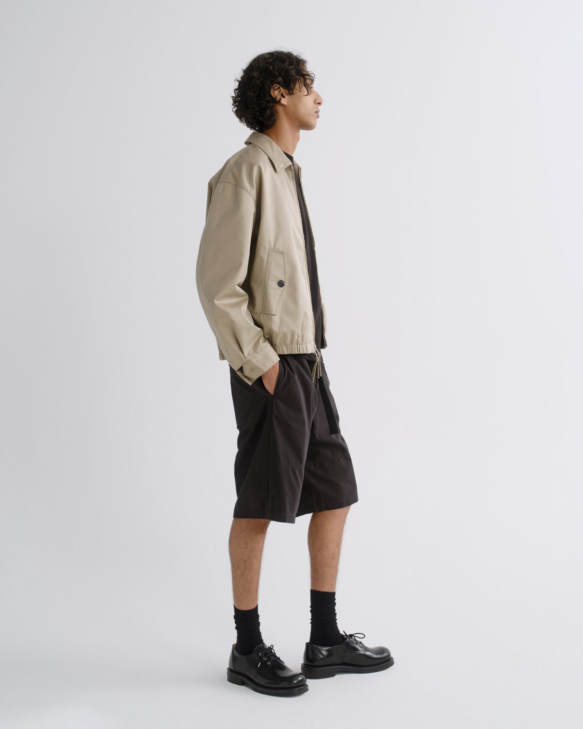 A model is shown in profile wearing a beige utility jacket, a black shirt, black shorts, black socks, and black leather shoes against a light gray studio background.