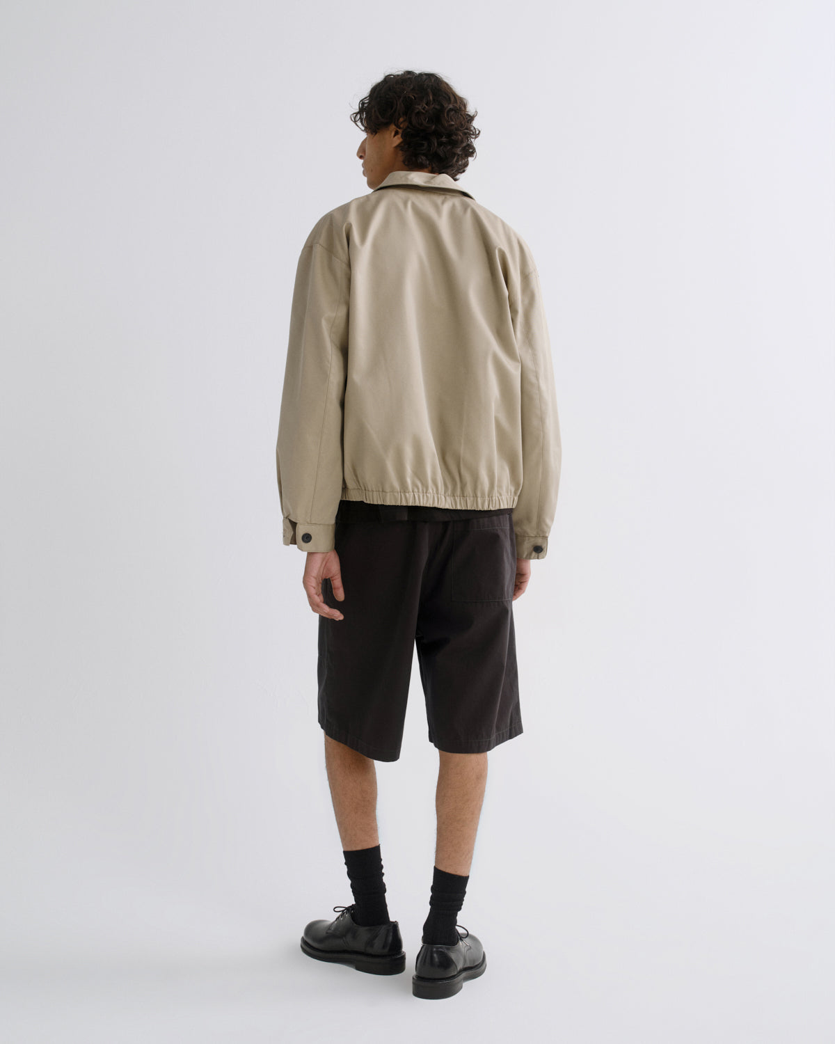 A model shown from the back wearing a beige utility jacket, black shorts, black socks, and black shoes, photographed against a plain light gray studio background.