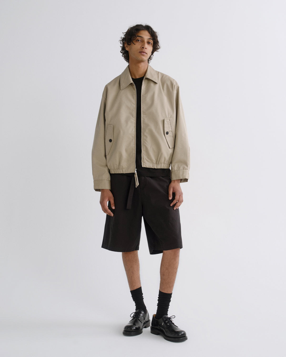 A model stands against a light gray studio background wearing a beige utility jacket over a black shirt, black shorts, black socks, and black leather shoes.