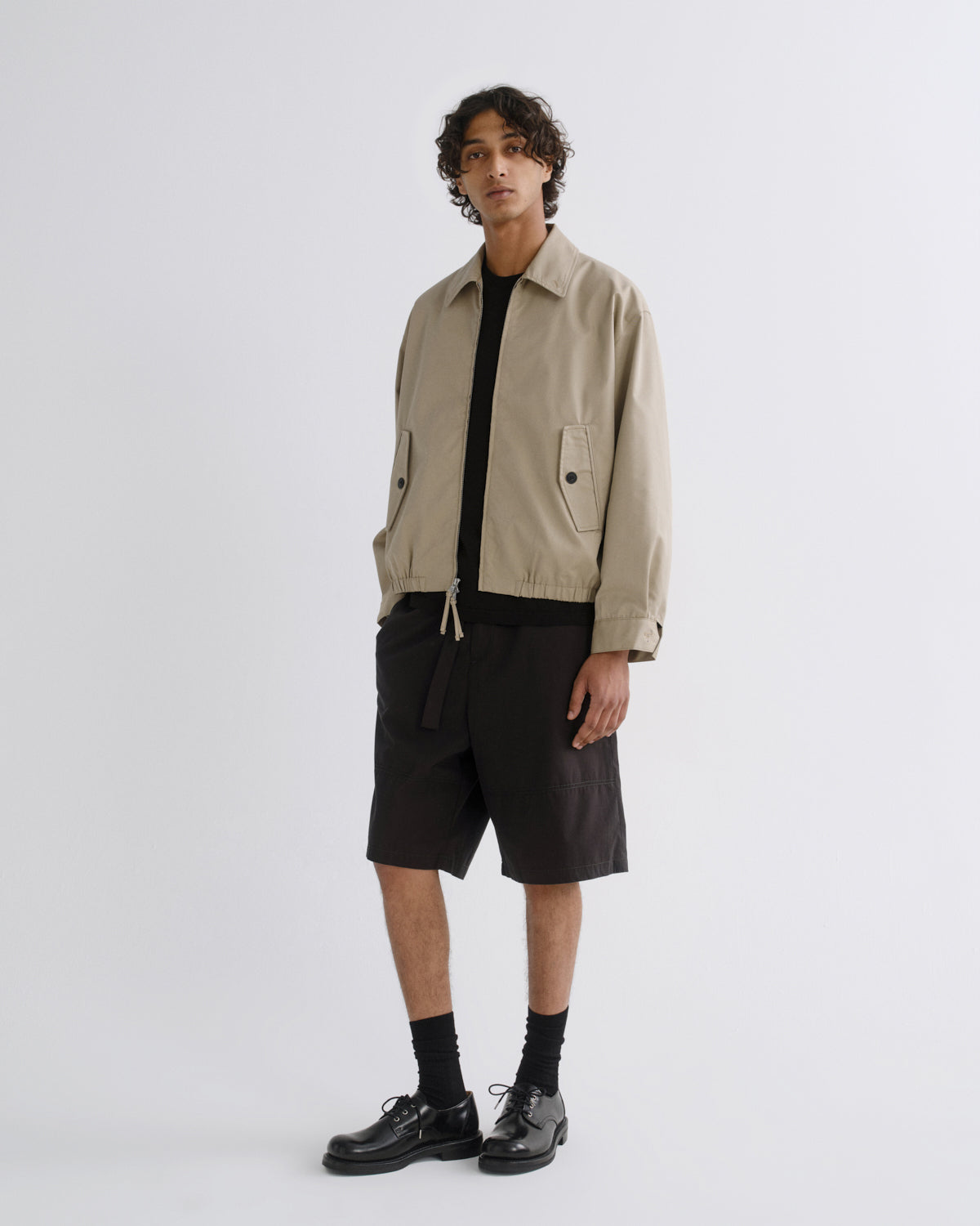 A model stands against a light gray studio background wearing a beige utility jacket over a black shirt, black shorts, black socks, and black leather shoes.