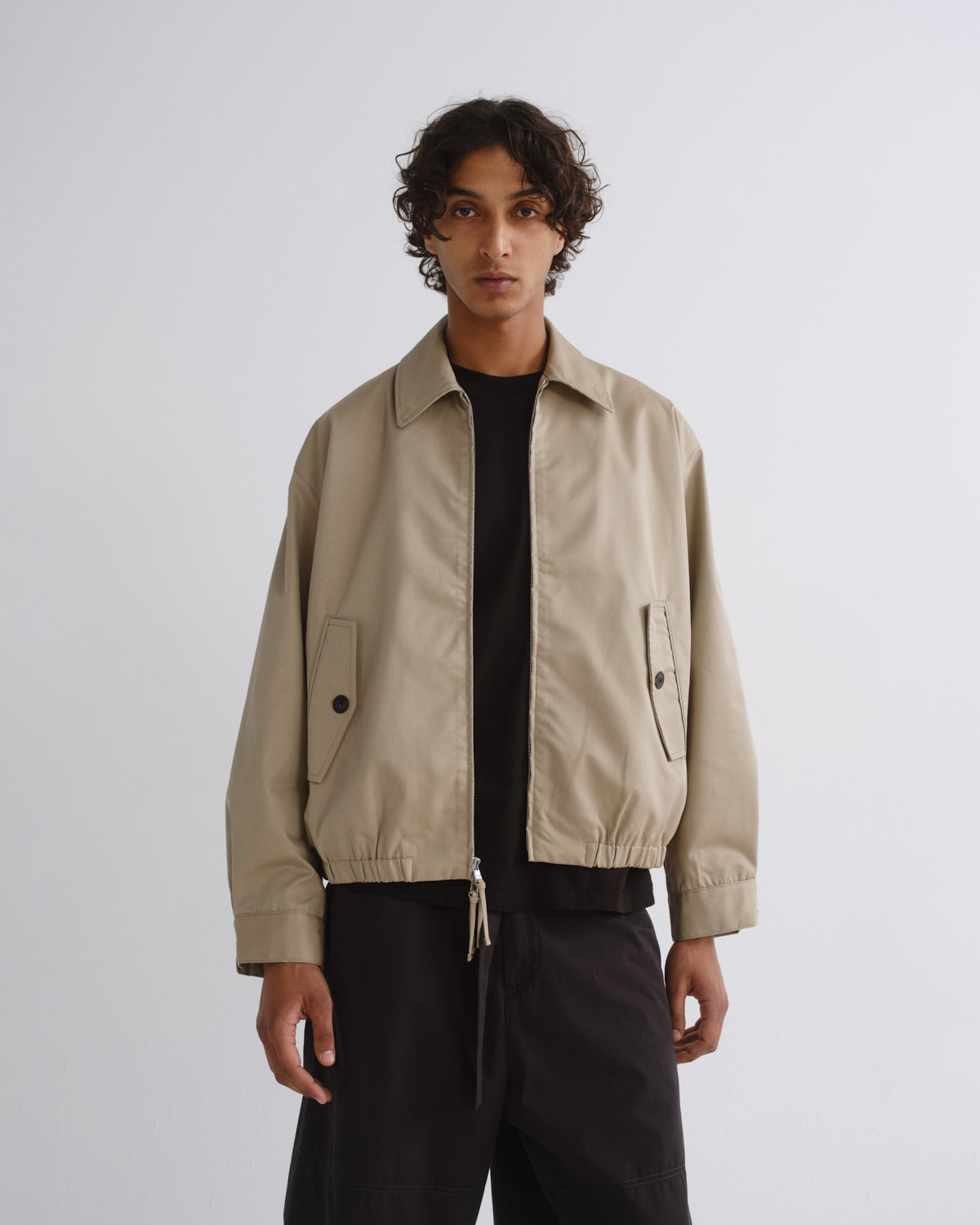 A man wearing a beige utility-style jacket over a black shirt and black pants, standing against a plain light gray studio background.