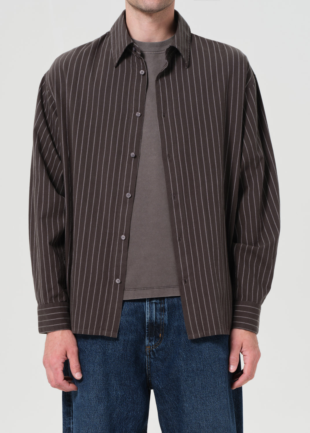 Man wearing a brown pinstripe shirt over a gray shirt with blue jeans on a white background