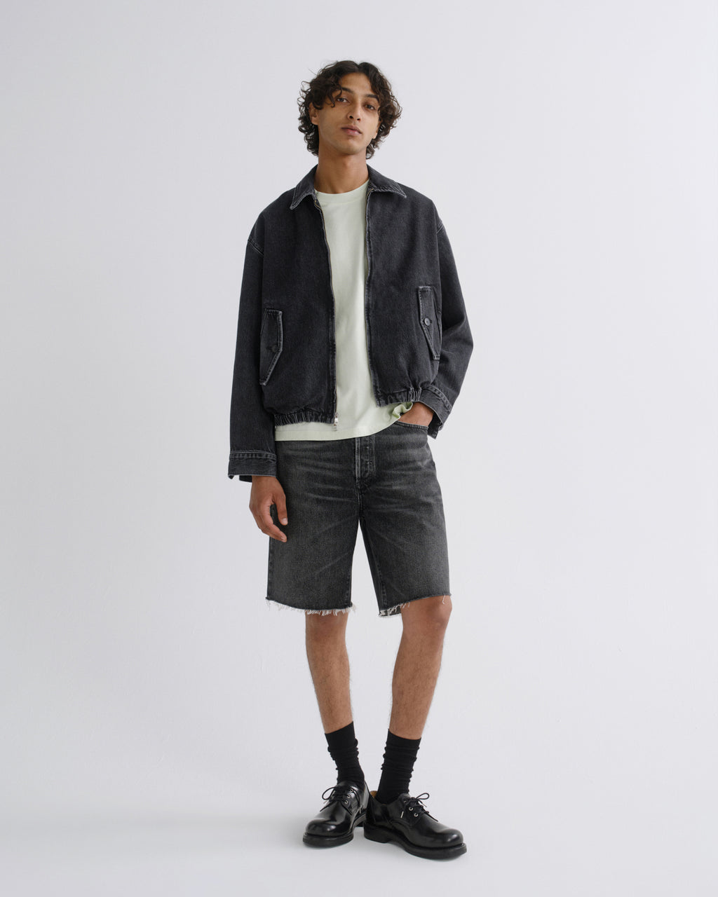 A model stands against a light gray studio background wearing a dark washed denim jacket over a white t-shirt, dark gray denim shorts, black socks, and black leather shoes.