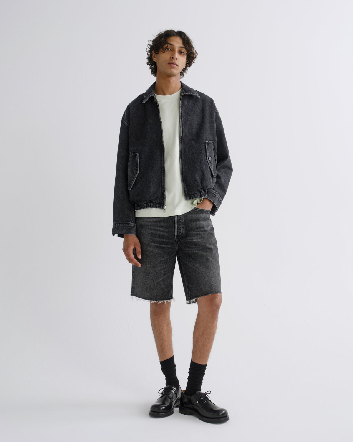 A model stands against a light gray studio background wearing a dark washed denim jacket over a white t-shirt, dark gray denim shorts, black socks, and black leather shoes.