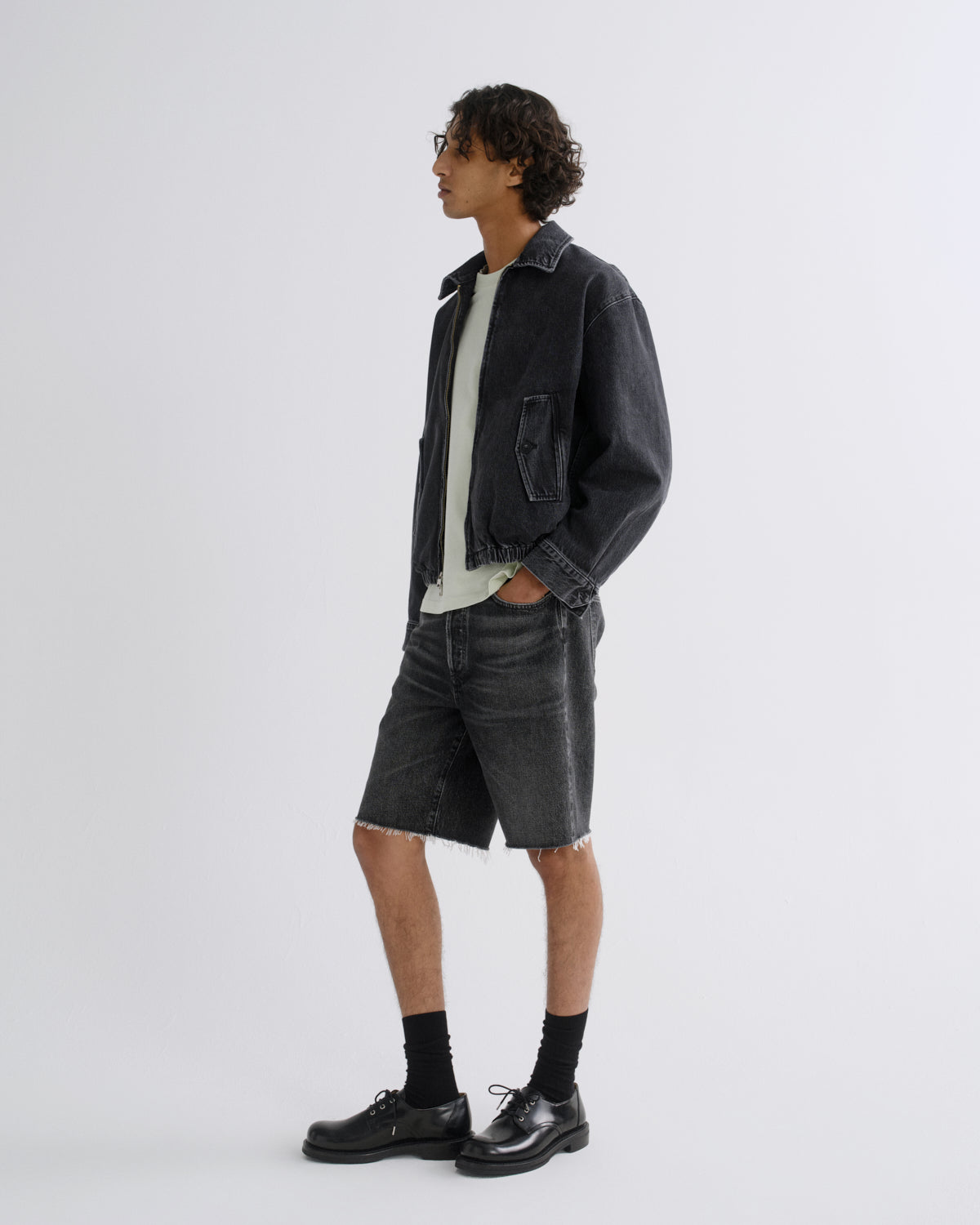 A model stands in profile wearing a dark washed denim jacket, matching denim shorts, a light gray t-shirt, black socks, and black leather shoes against a plain light gray studio background.