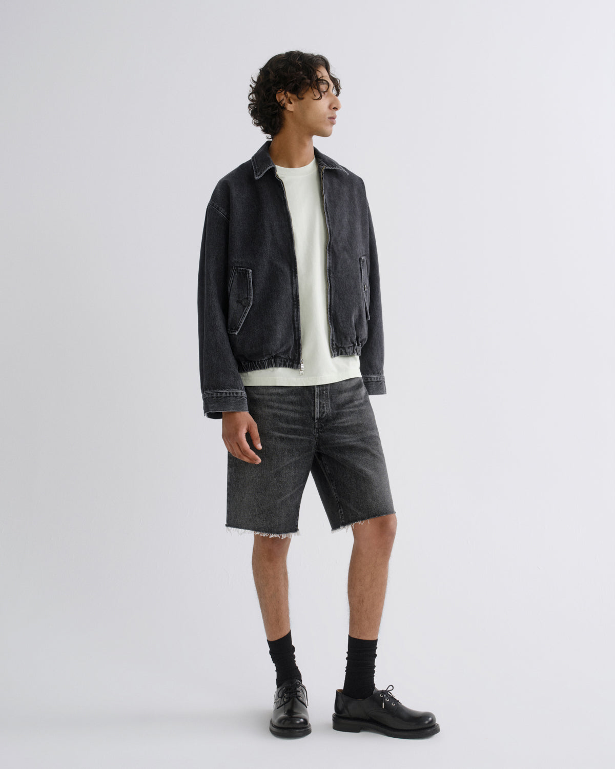 A model stands in profile wearing a dark washed denim jacket over a white t-shirt, dark denim shorts, black socks, and black leather shoes, photographed against a light gray studio background.