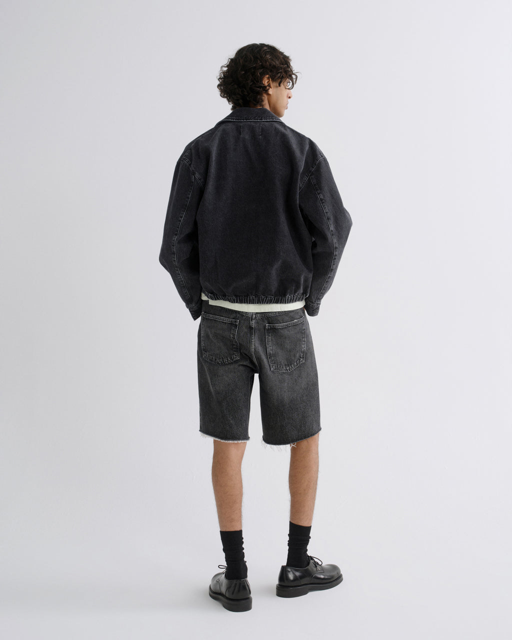 A model shown from the back wearing a washed black denim jacket, faded black denim shorts, black socks, and black leather shoes, photographed against a light gray studio background.