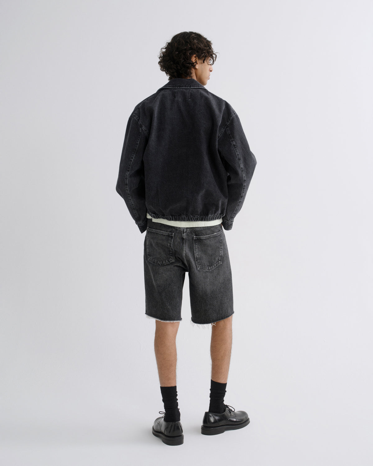 A model shown from the back wearing a washed black denim jacket, faded black denim shorts, black socks, and black leather shoes, photographed against a light gray studio background.