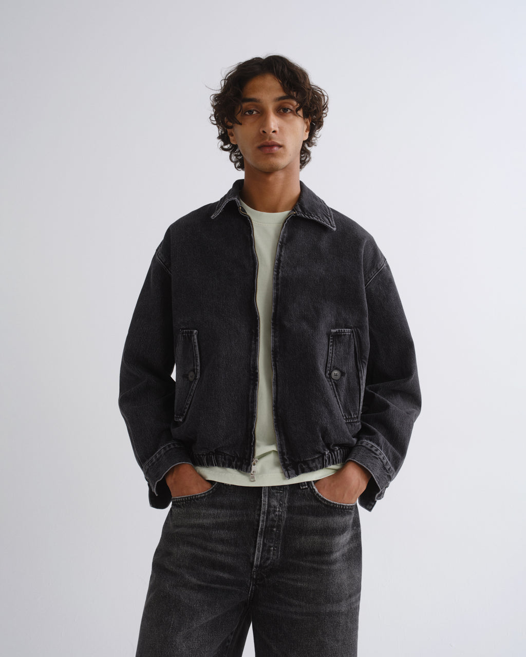 A man wearing a dark washed denim jacket over a light green t-shirt and dark jeans, standing against a light gray studio background.