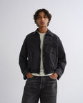 A man wearing a dark washed denim jacket over a light green t-shirt and dark jeans, standing against a light gray studio background.