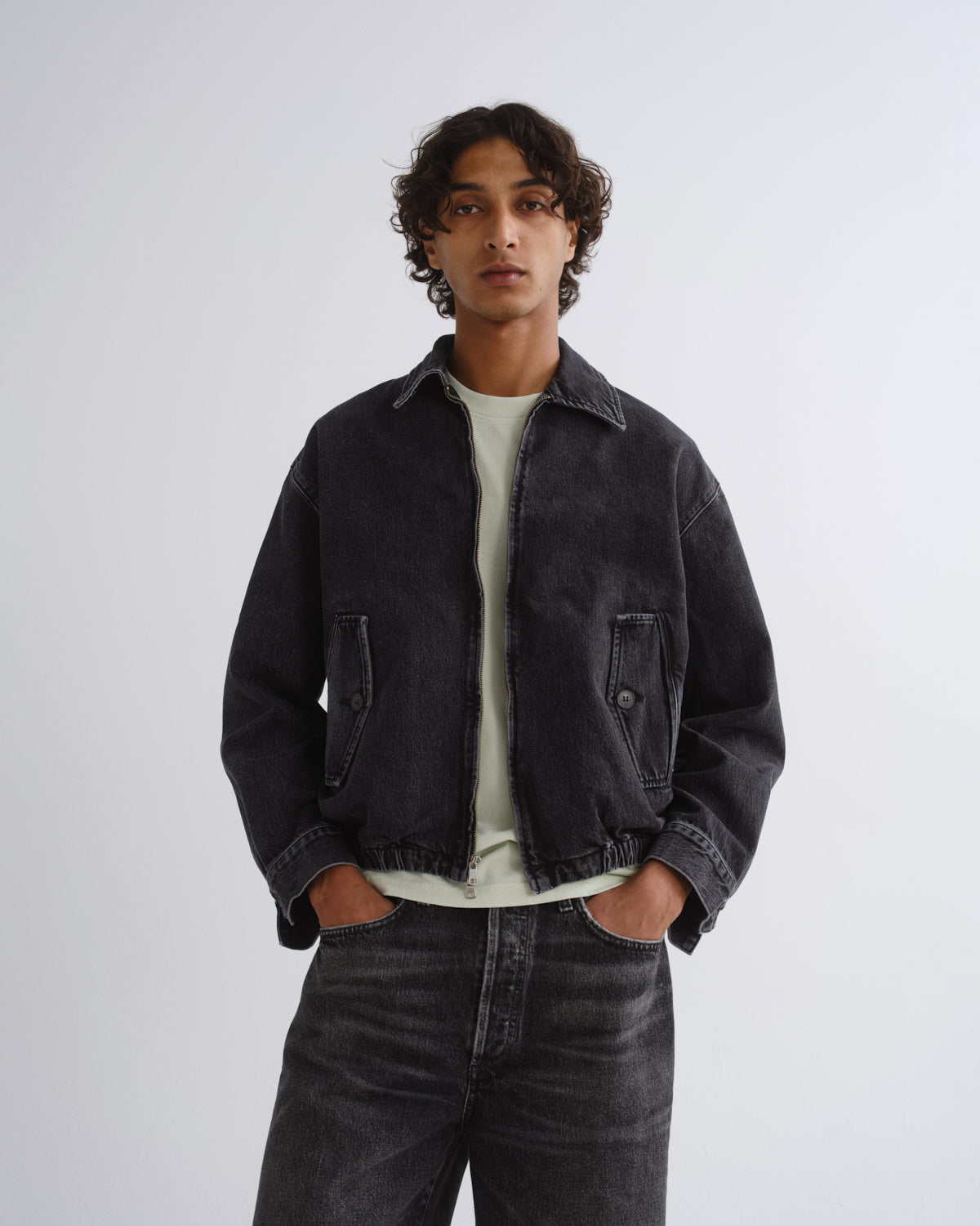 A man wearing a dark washed denim jacket over a light green t-shirt and dark jeans, standing against a light gray studio background.