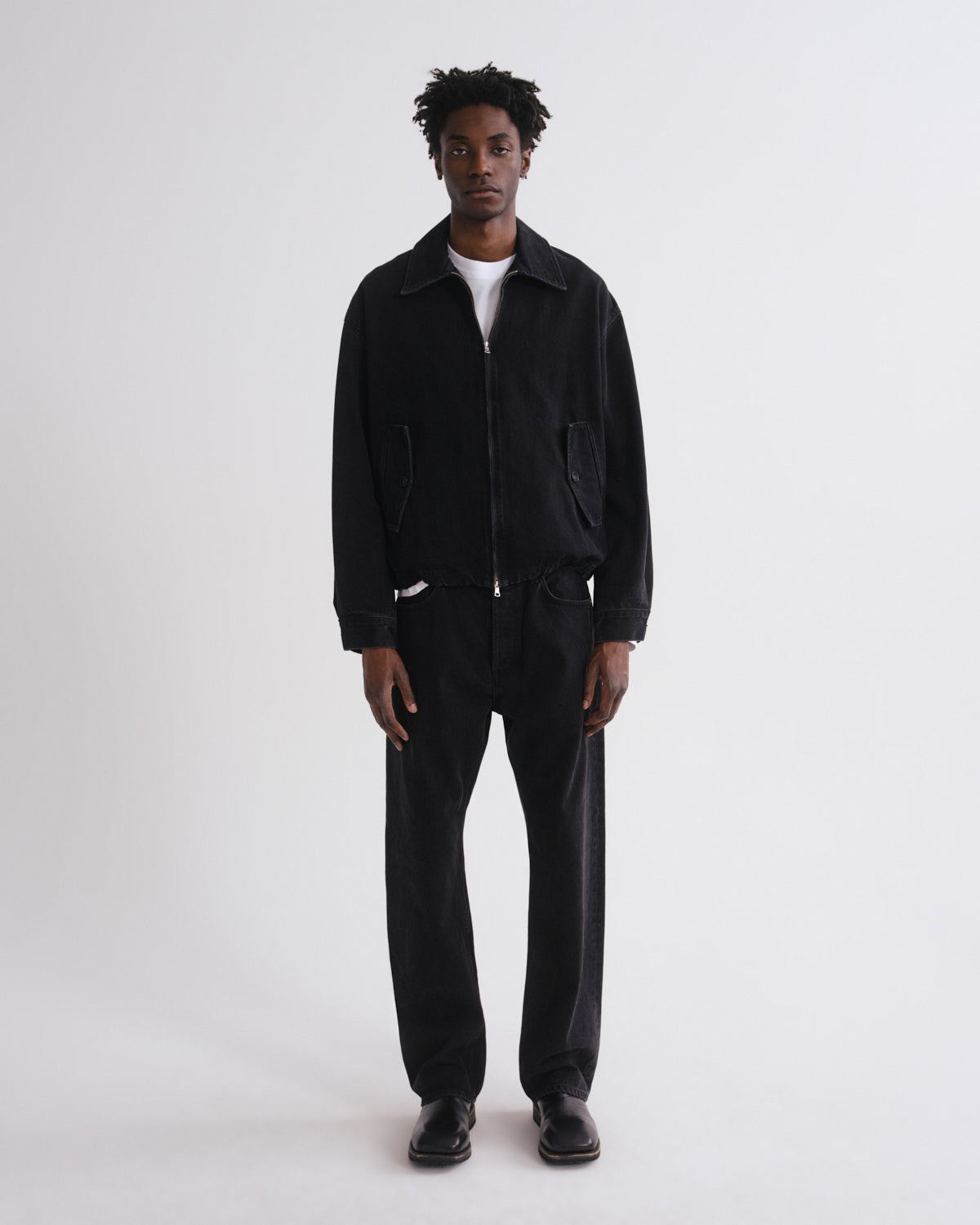 A man stands against a plain light gray studio background wearing a black zip-front jacket with large front pockets, black pants, black shoes, and a white shirt underneath.