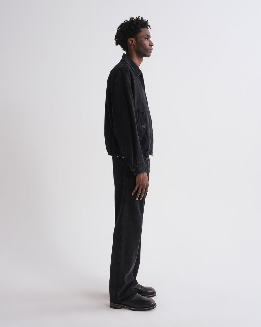 A man in a black denim jacket and black wide-leg jeans stands in profile against a plain light gray studio background.