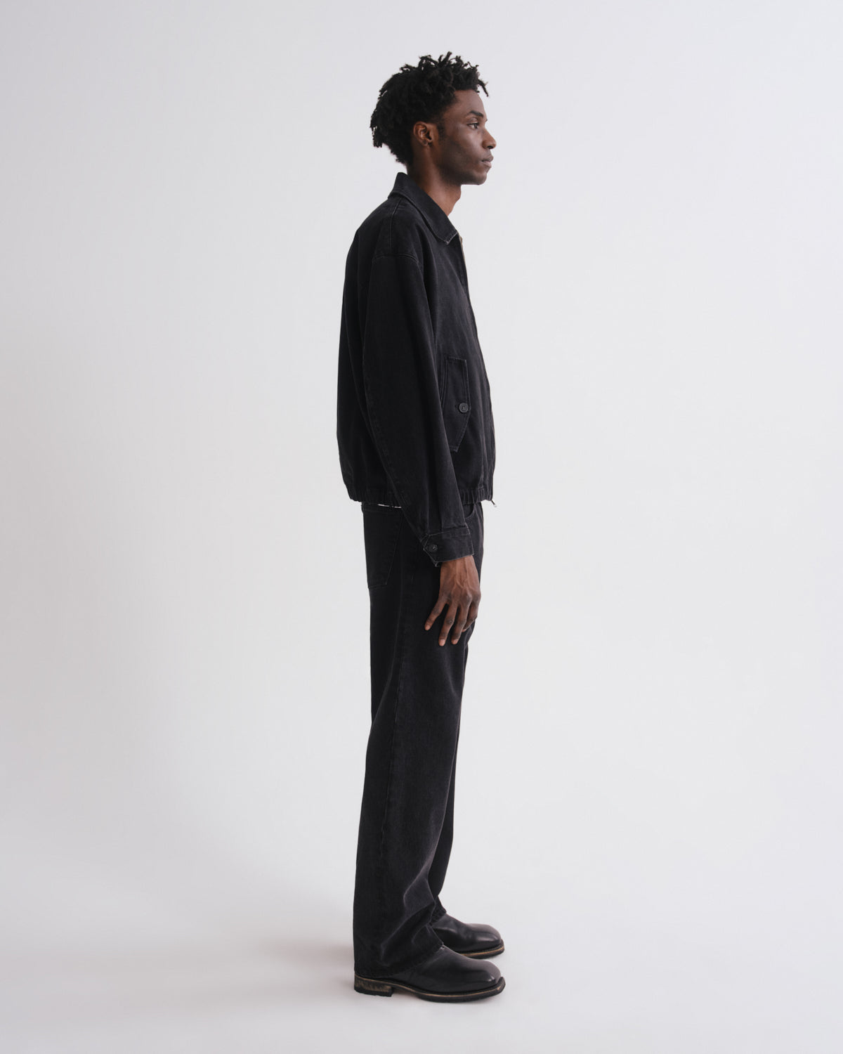 A man in a black denim jacket and black wide-leg jeans stands in profile against a plain light gray studio background.