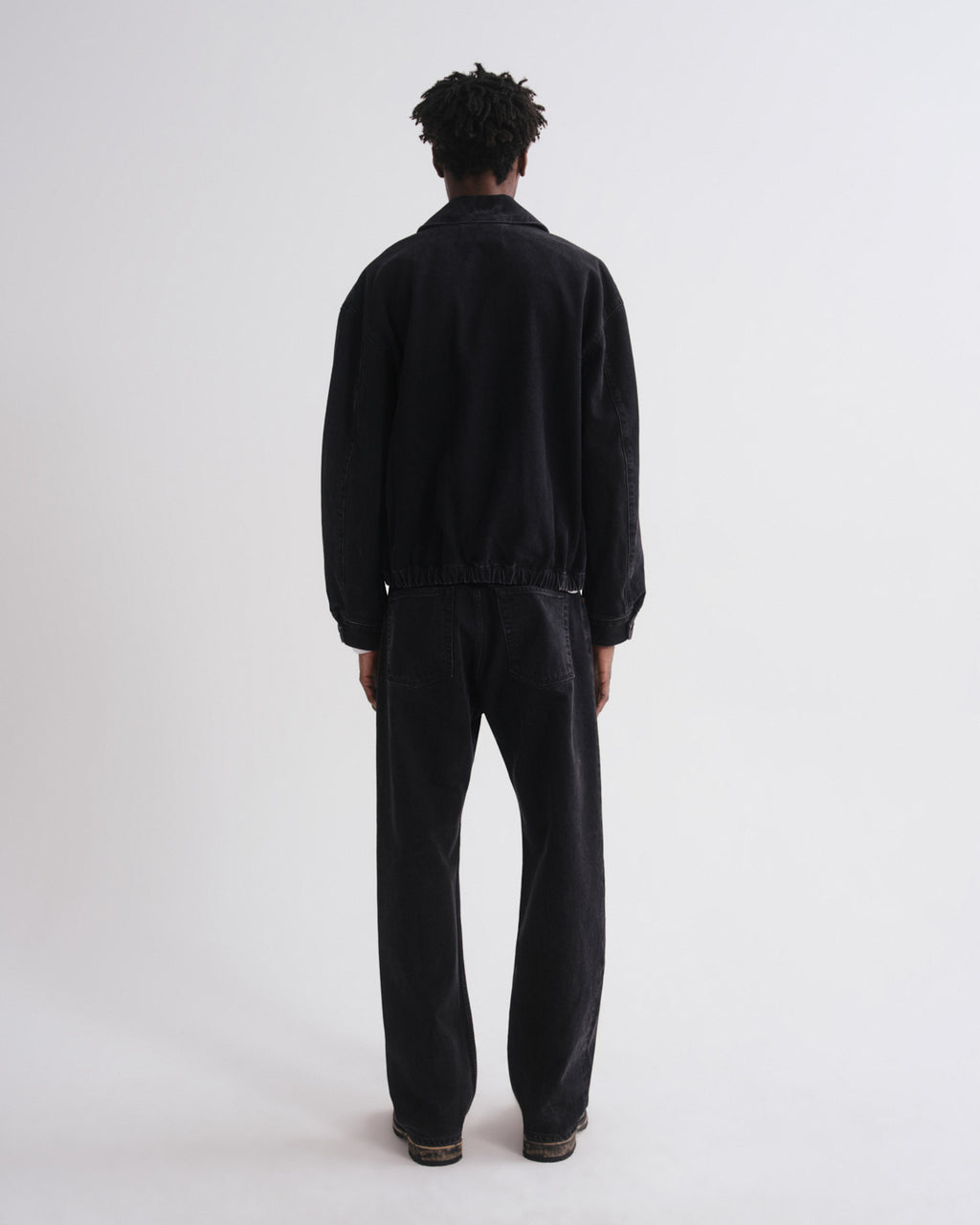 A model viewed from the back wearing a black jacket and matching black trousers against a plain light gray studio background.