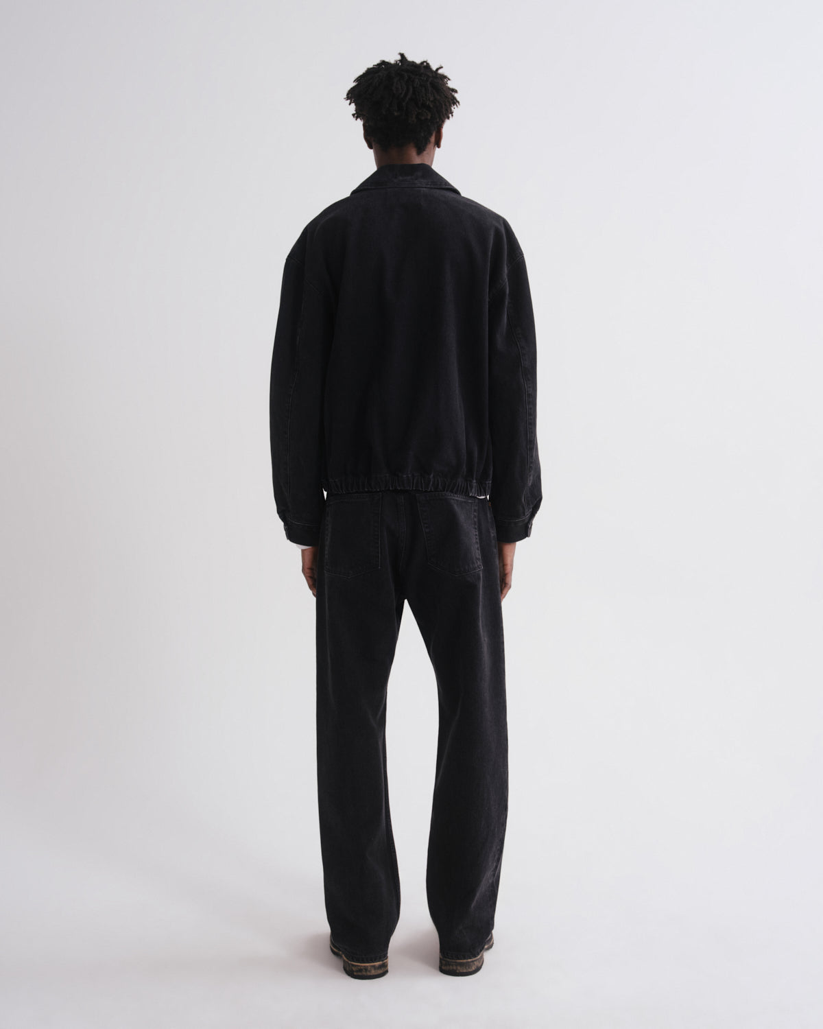 A model viewed from the back wearing a black jacket and matching black trousers against a plain light gray studio background.