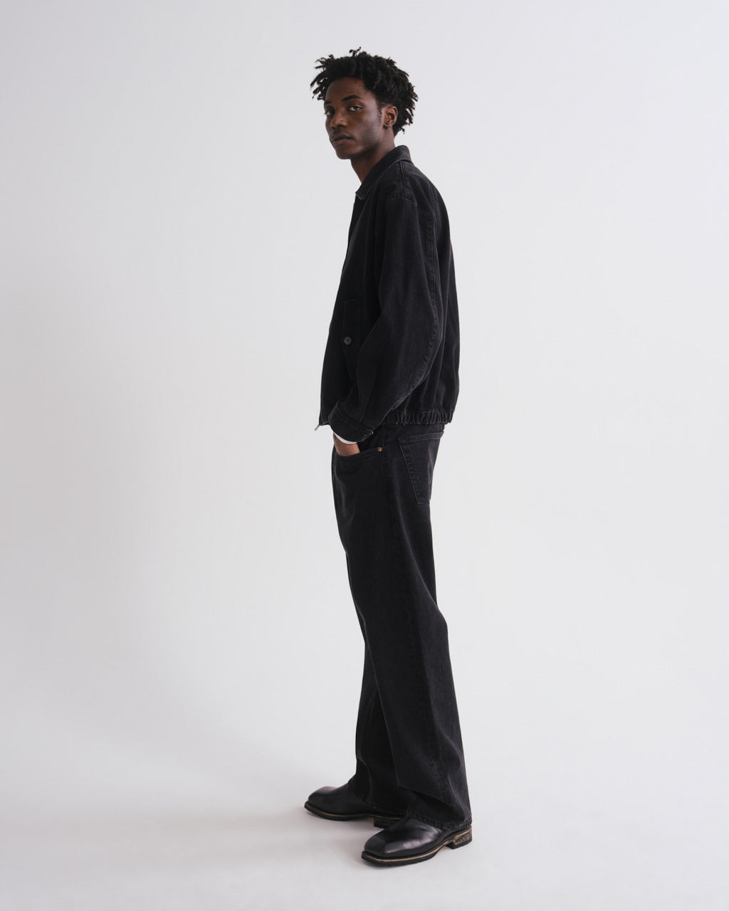 A man stands in profile wearing a black corduroy jacket, black wide-leg jeans, and black leather shoes against a plain light gray studio background.