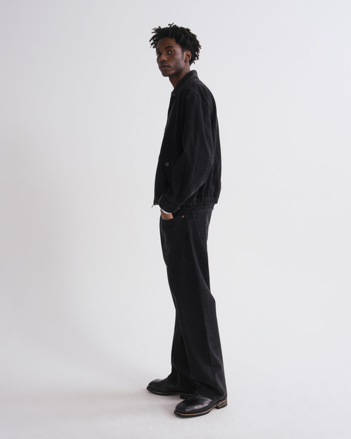 A man stands in profile wearing a black corduroy jacket, black wide-leg jeans, and black leather shoes against a plain light gray studio background.