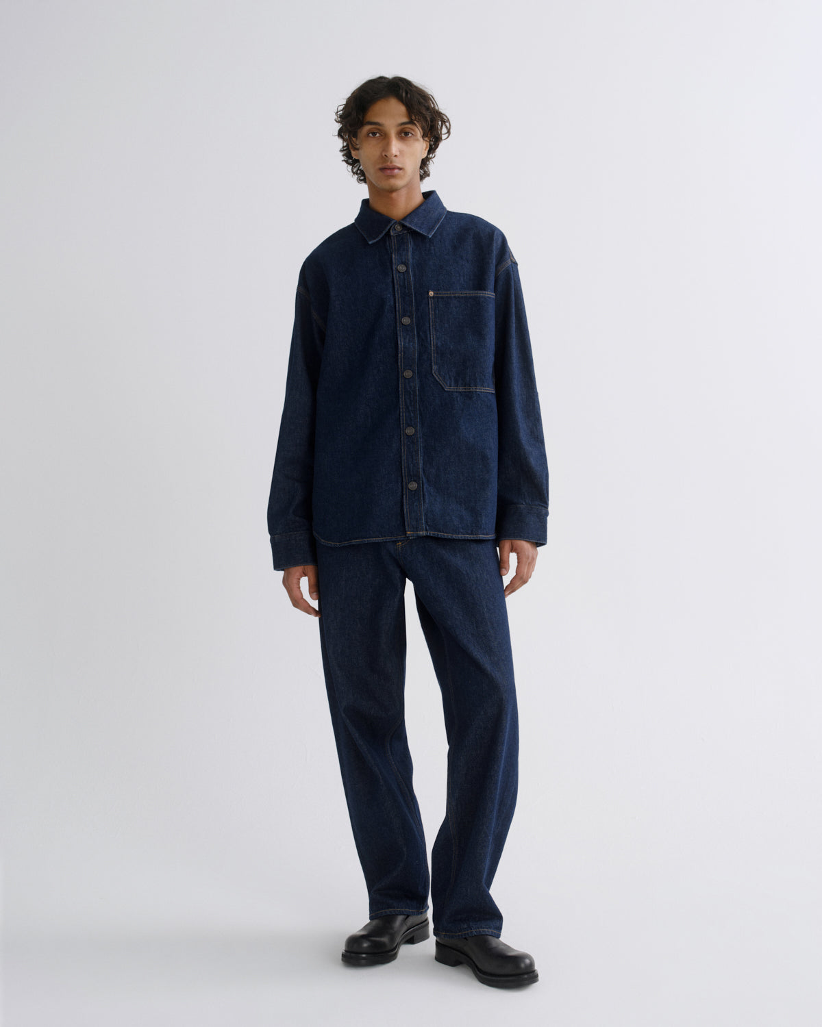 A model stands against a light gray studio background wearing a dark indigo denim long-sleeve button-up shirt and matching dark indigo denim trousers, paired with black leather shoes.