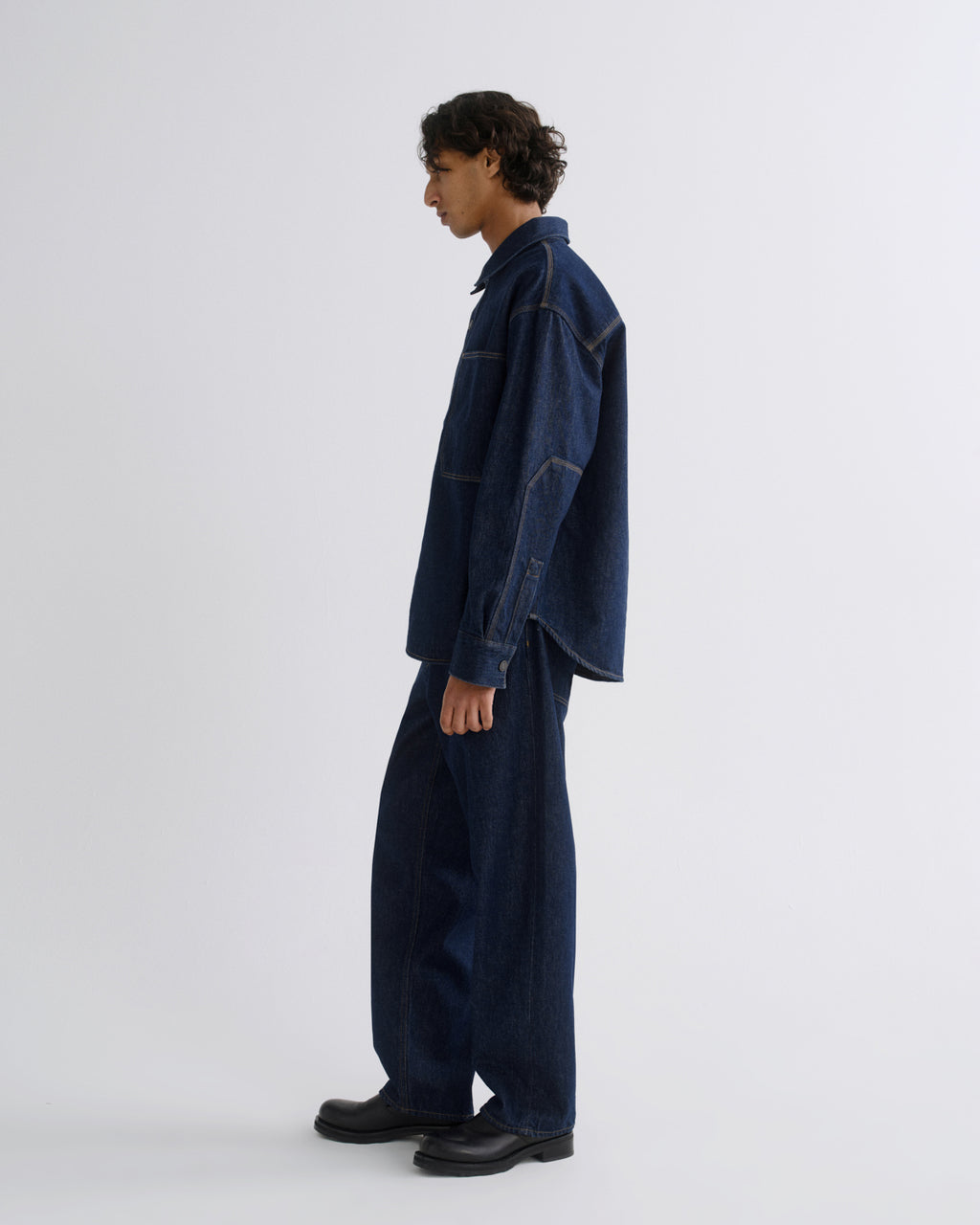 A model in side profile wearing a dark indigo denim jacket and matching wide-leg denim pants, paired with black leather shoes, against a plain light gray studio background.