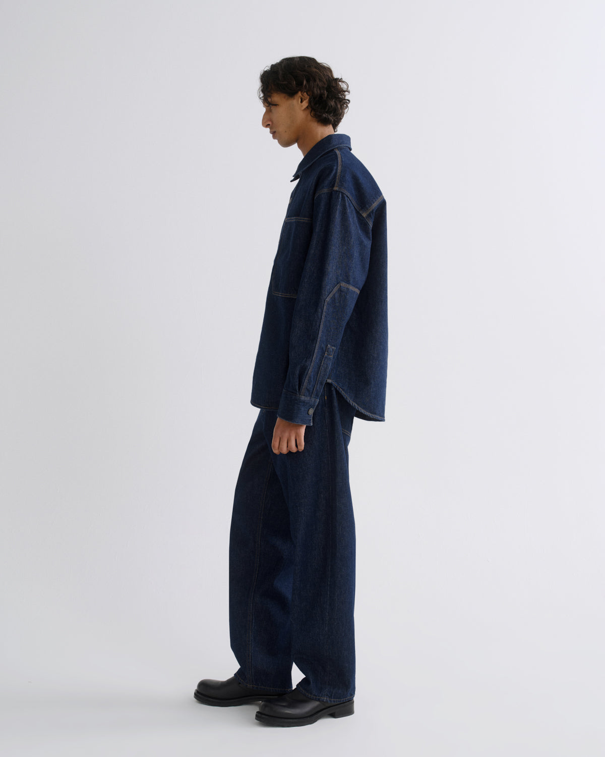 A model in side profile wearing a dark indigo denim jacket and matching wide-leg denim pants, paired with black leather shoes, against a plain light gray studio background.