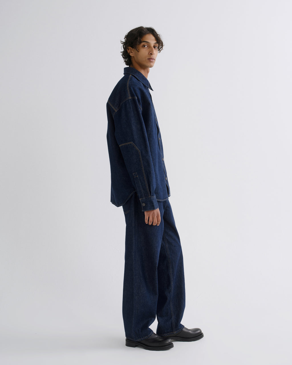 A model wearing a dark indigo denim work jacket and matching wide-leg trousers, standing in profile against a light gray studio background.