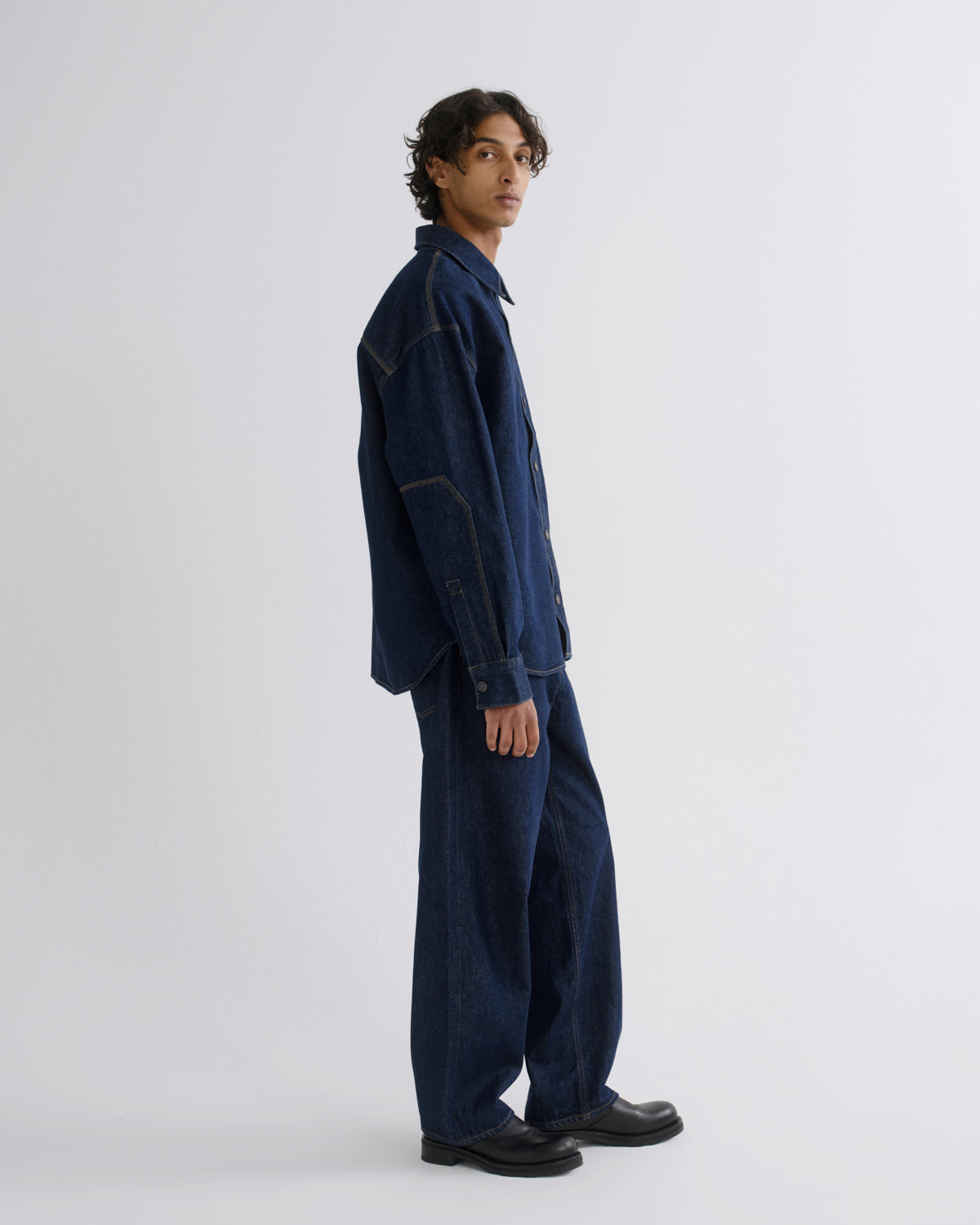 A model wearing a dark indigo denim work jacket and matching wide-leg trousers, standing in profile against a light gray studio background.