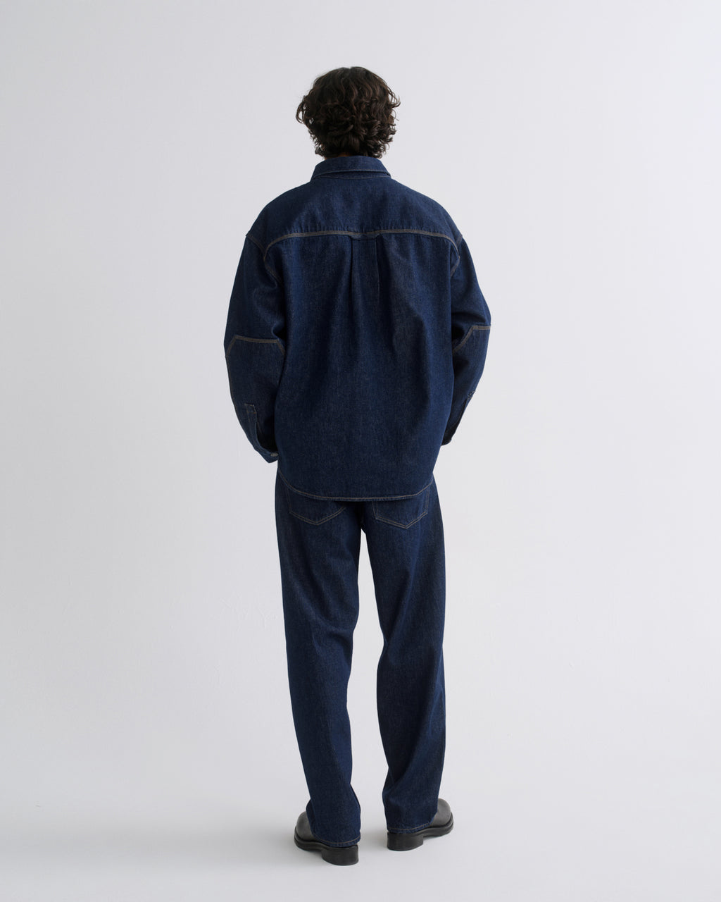 A man viewed from behind wearing a dark indigo denim work jacket and matching dark blue denim pants, standing against a light gray studio background.