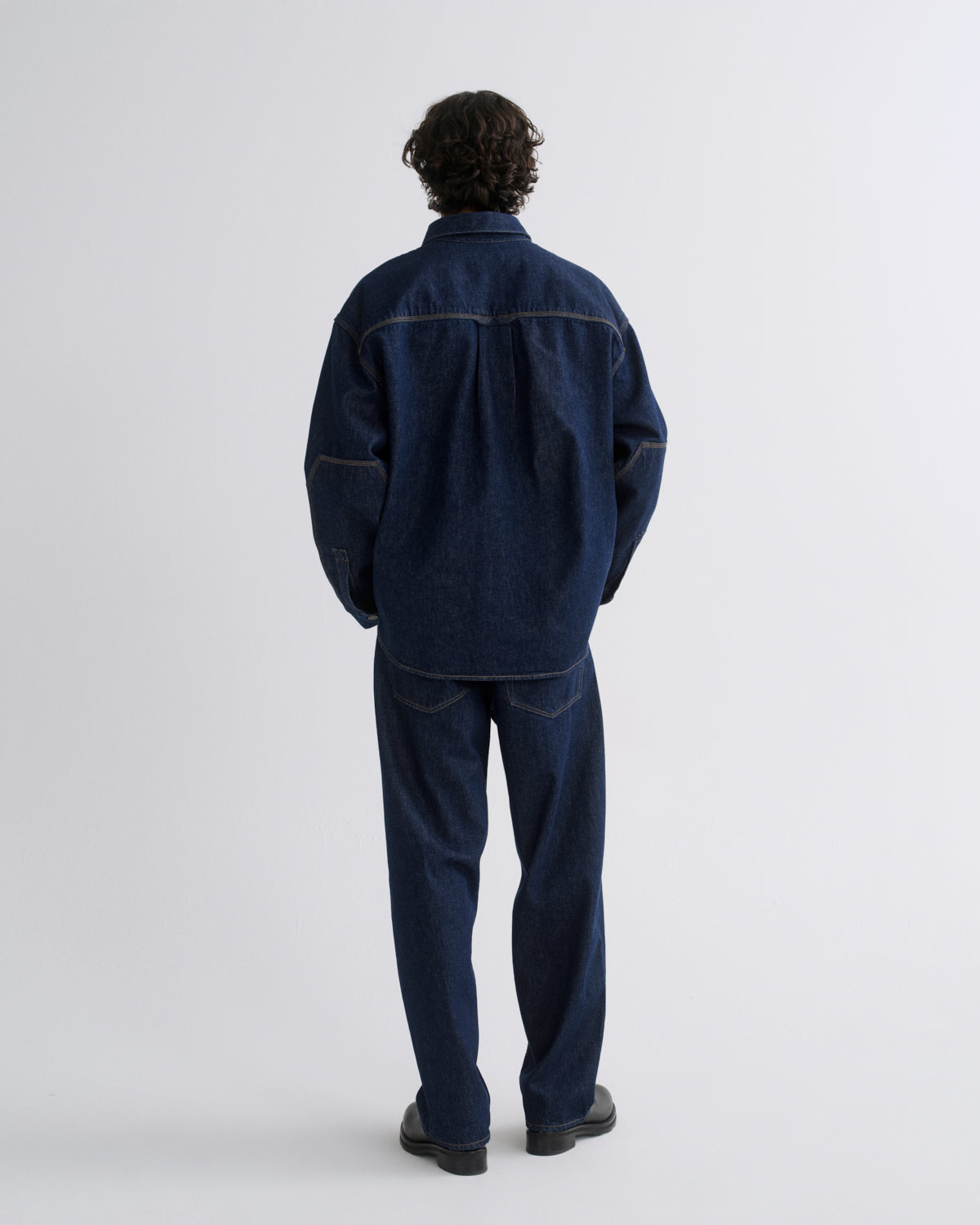 A man viewed from behind wearing a dark indigo denim work jacket and matching dark blue denim pants, standing against a light gray studio background.
