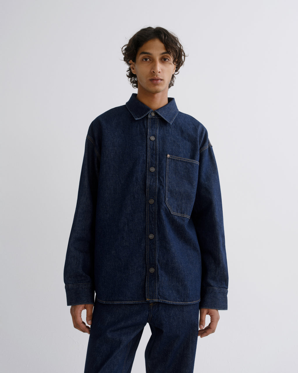 A man wearing a dark indigo denim long-sleeve button-up shirt and matching dark denim pants, standing against a light gray studio background.