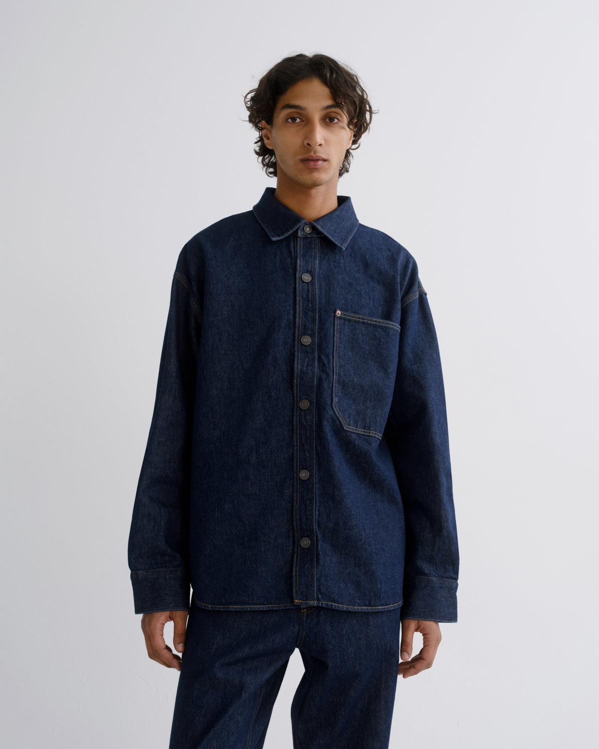 A man wearing a dark indigo denim long-sleeve button-up shirt and matching dark denim pants, standing against a light gray studio background.