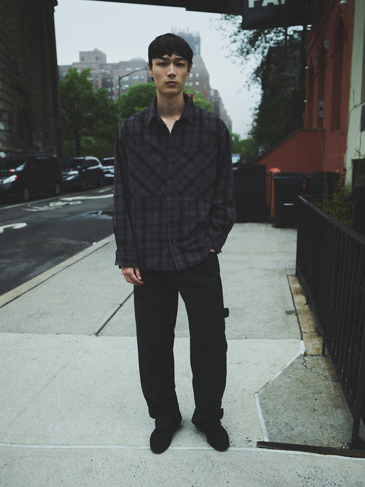Person standing on a sidewalk wearing a dark plaid shirt and black pants.