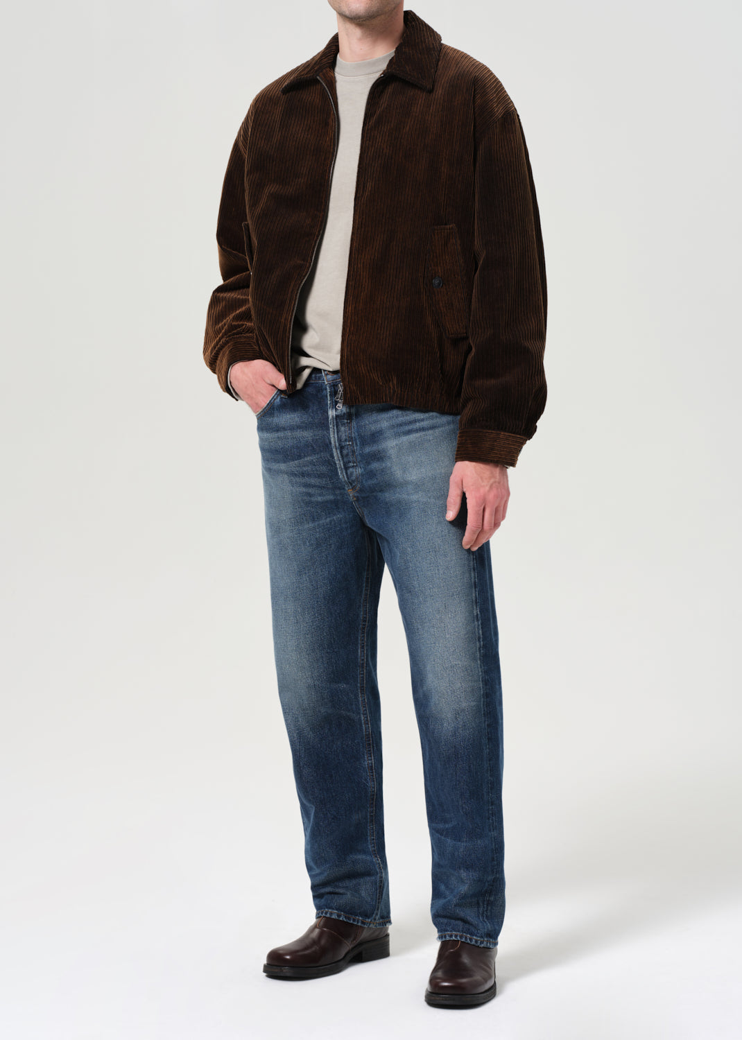 Person wearing a brown jacket, white shirt, blue jeans, and dark shoes on a white background