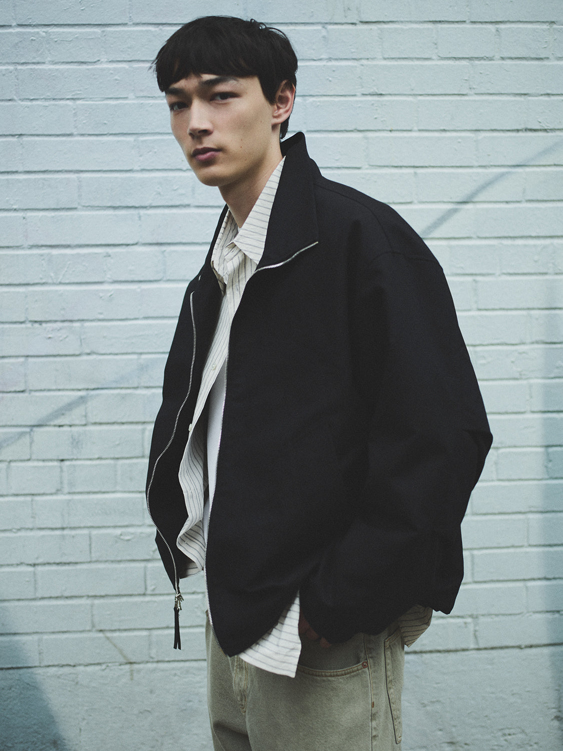 Man wearing a black jacket over a white shirt against a light blue brick wall.