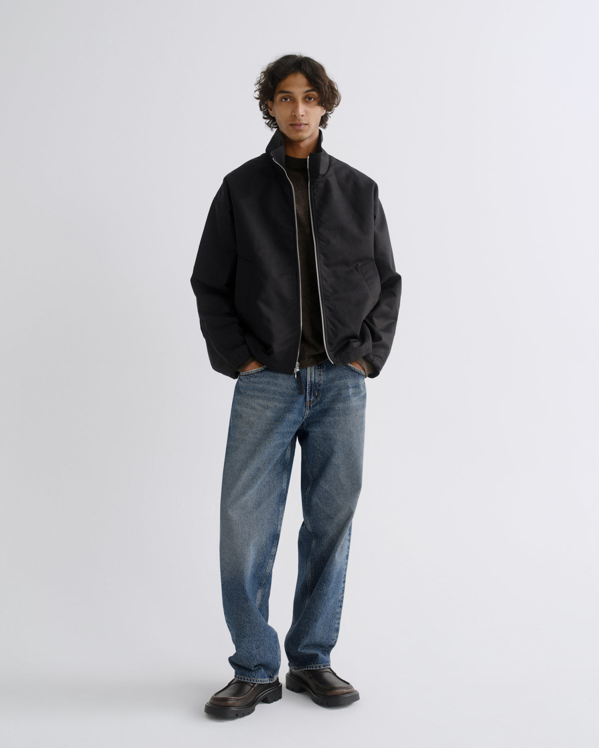 A model stands against a light gray studio background wearing a black zip-up jacket, blue jeans, and dark brown leather shoes.