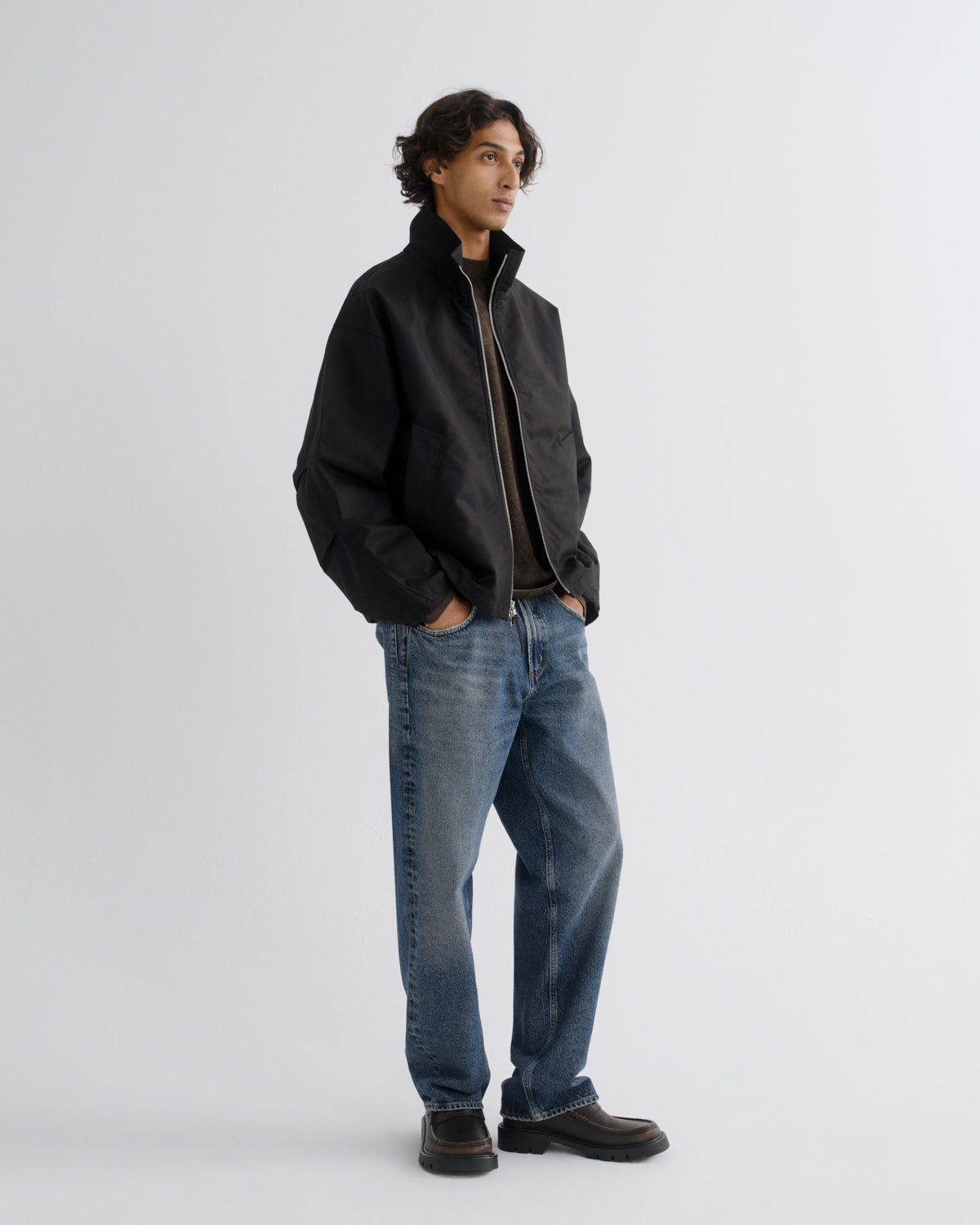 A man wearing a black zip-up jacket, blue jeans, and dark brown shoes, standing against a light gray studio background.