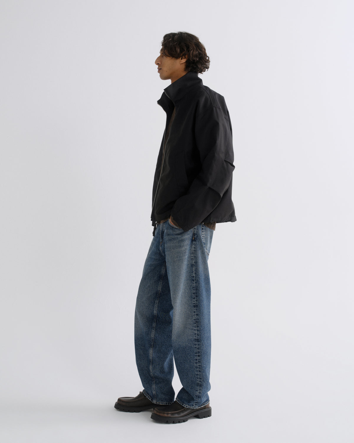 A man in profile wearing a black jacket, blue jeans, and dark brown shoes, standing against a plain light gray studio background.