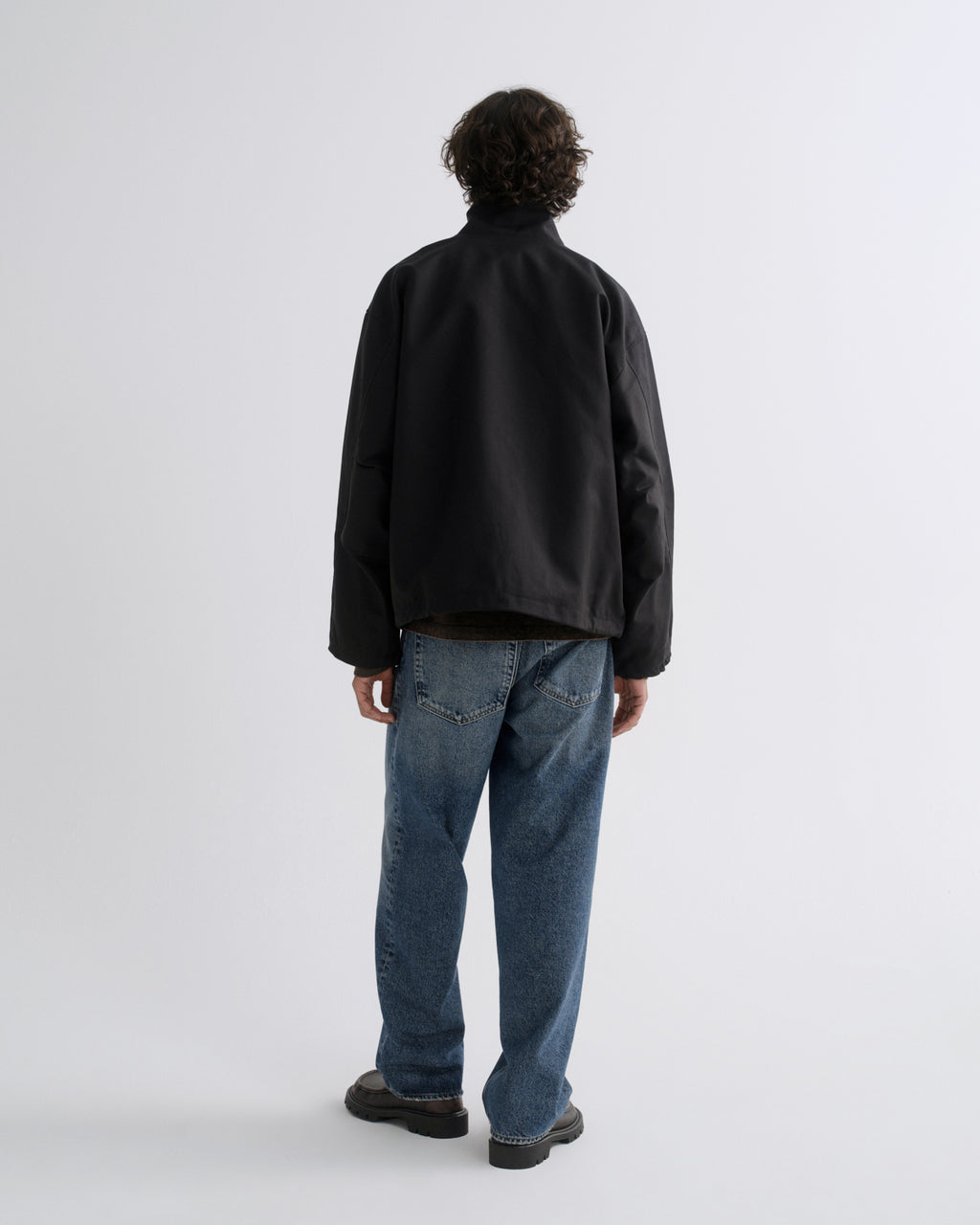 A man viewed from behind wearing a black jacket, blue jeans, and dark brown shoes, standing against a light gray studio background.