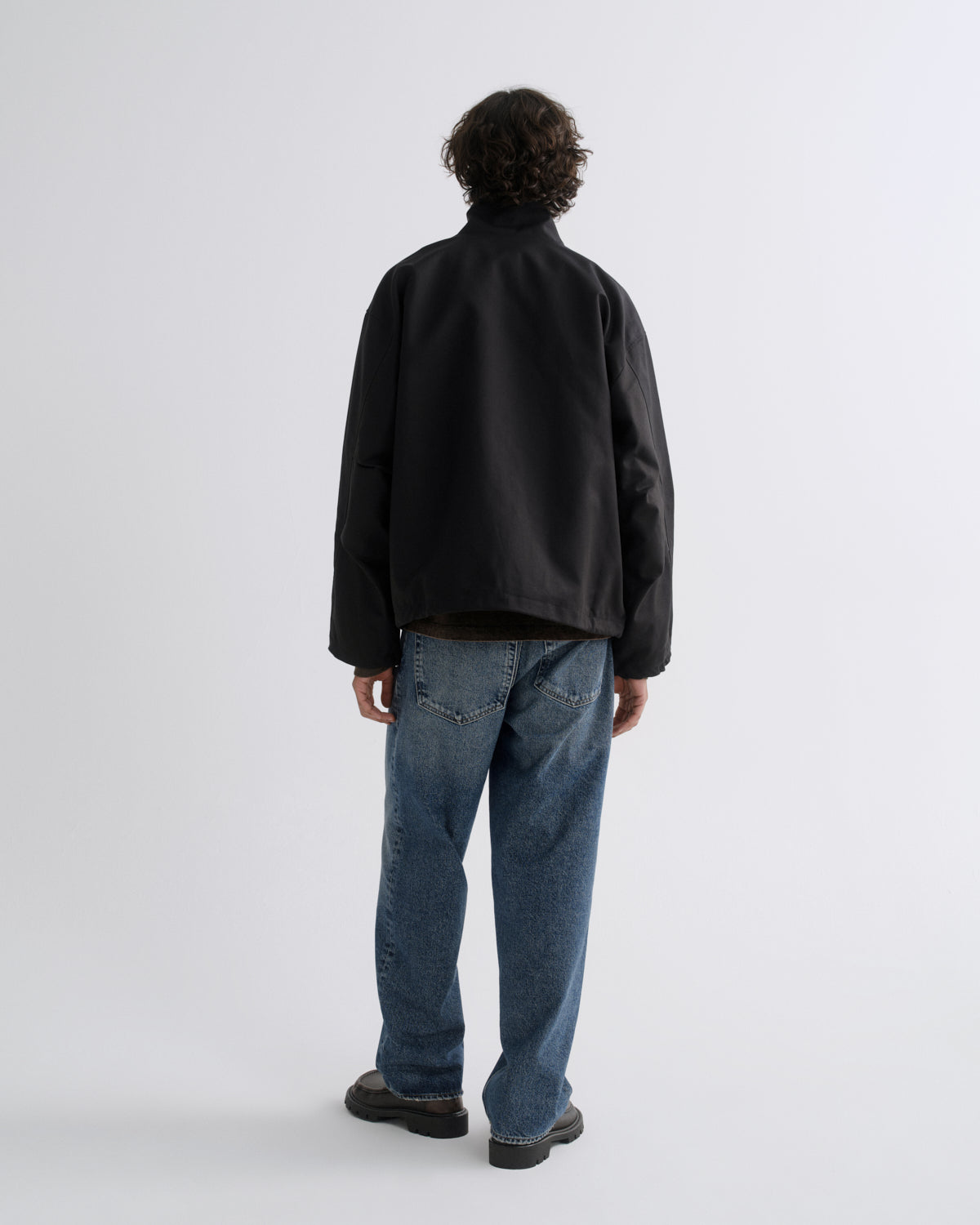 A man viewed from behind wearing a black jacket, blue jeans, and dark brown shoes, standing against a light gray studio background.