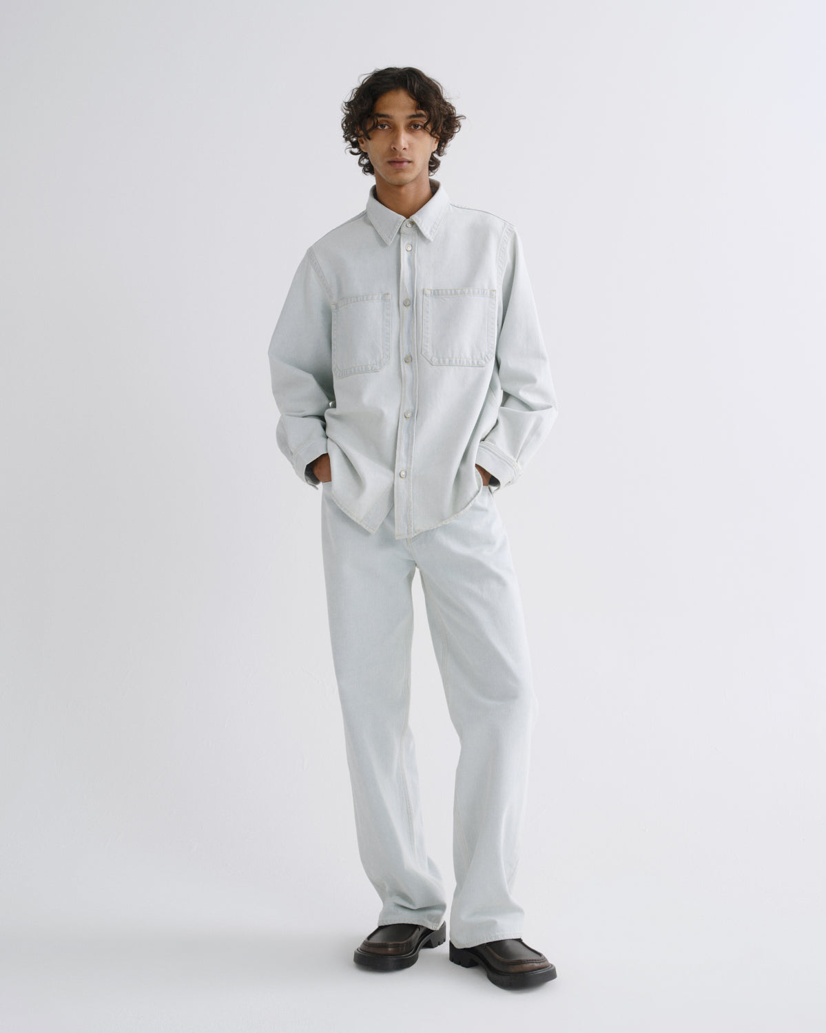 A model stands against a light gray studio background wearing a light gray long-sleeve button-up shirt and matching light gray trousers, paired with dark brown loafers.