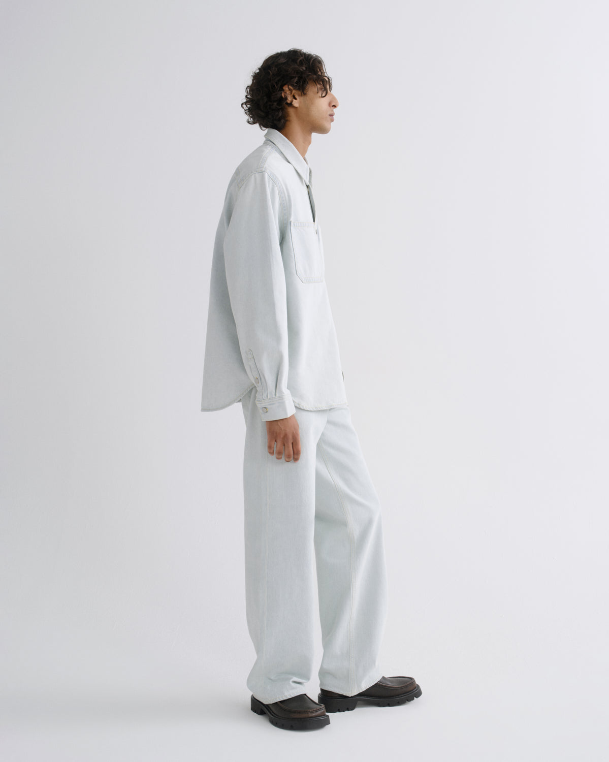 A model shown in profile wearing a light blue oversized button-up shirt and matching wide-leg trousers, paired with dark brown shoes, against a plain light gray studio background.