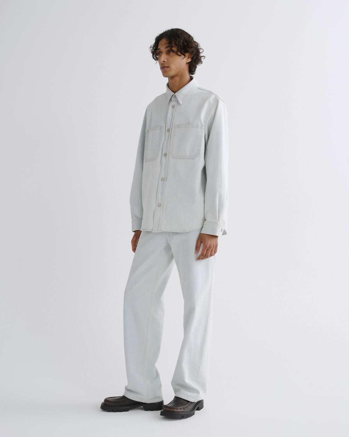 A model stands in a studio wearing a light gray long-sleeve button-up shirt and matching light gray trousers, paired with dark brown shoes, against a plain light gray background.