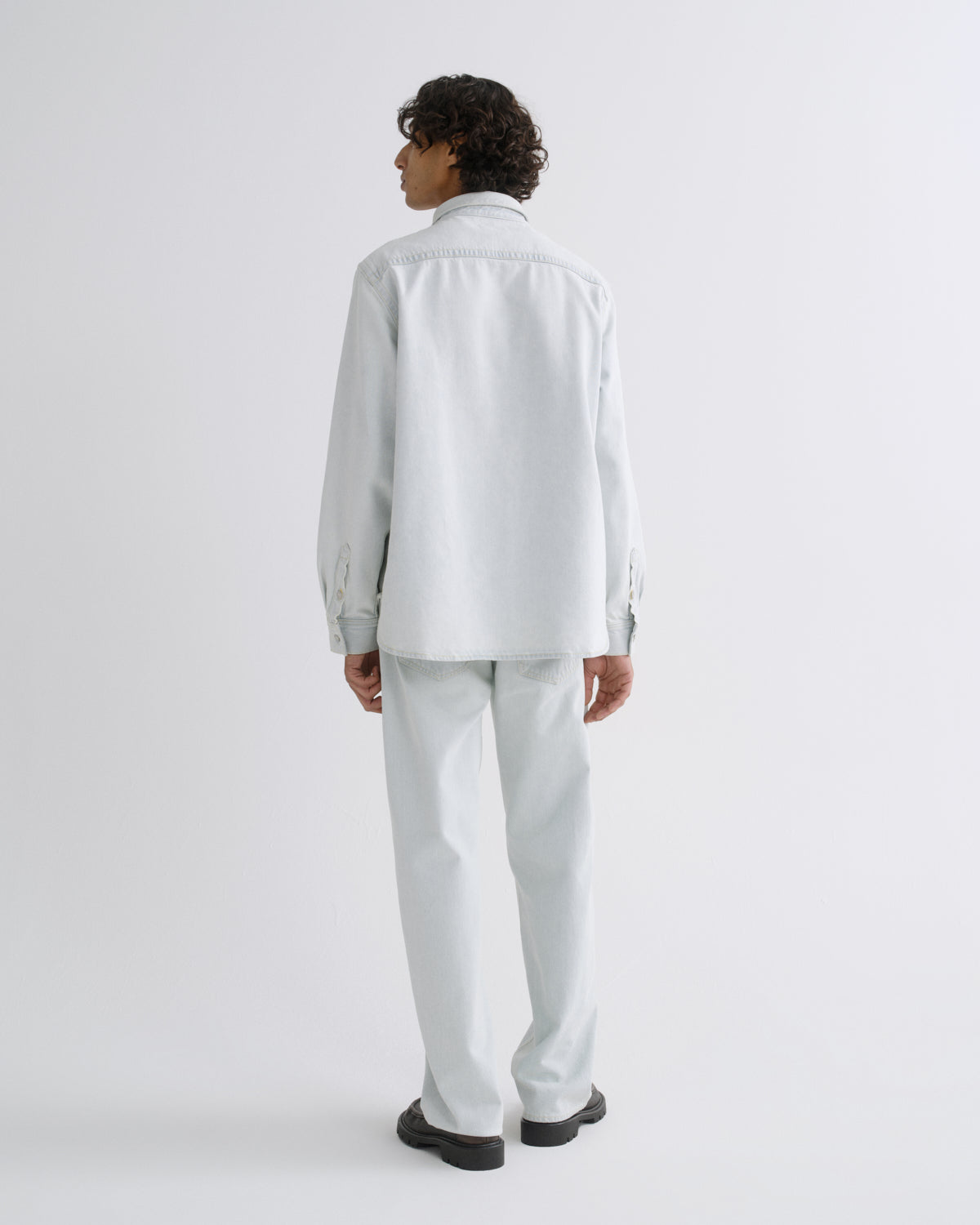 A model shown from the back wearing a light gray long-sleeve button-up shirt and matching light gray trousers, paired with black shoes, against a plain light gray studio background.