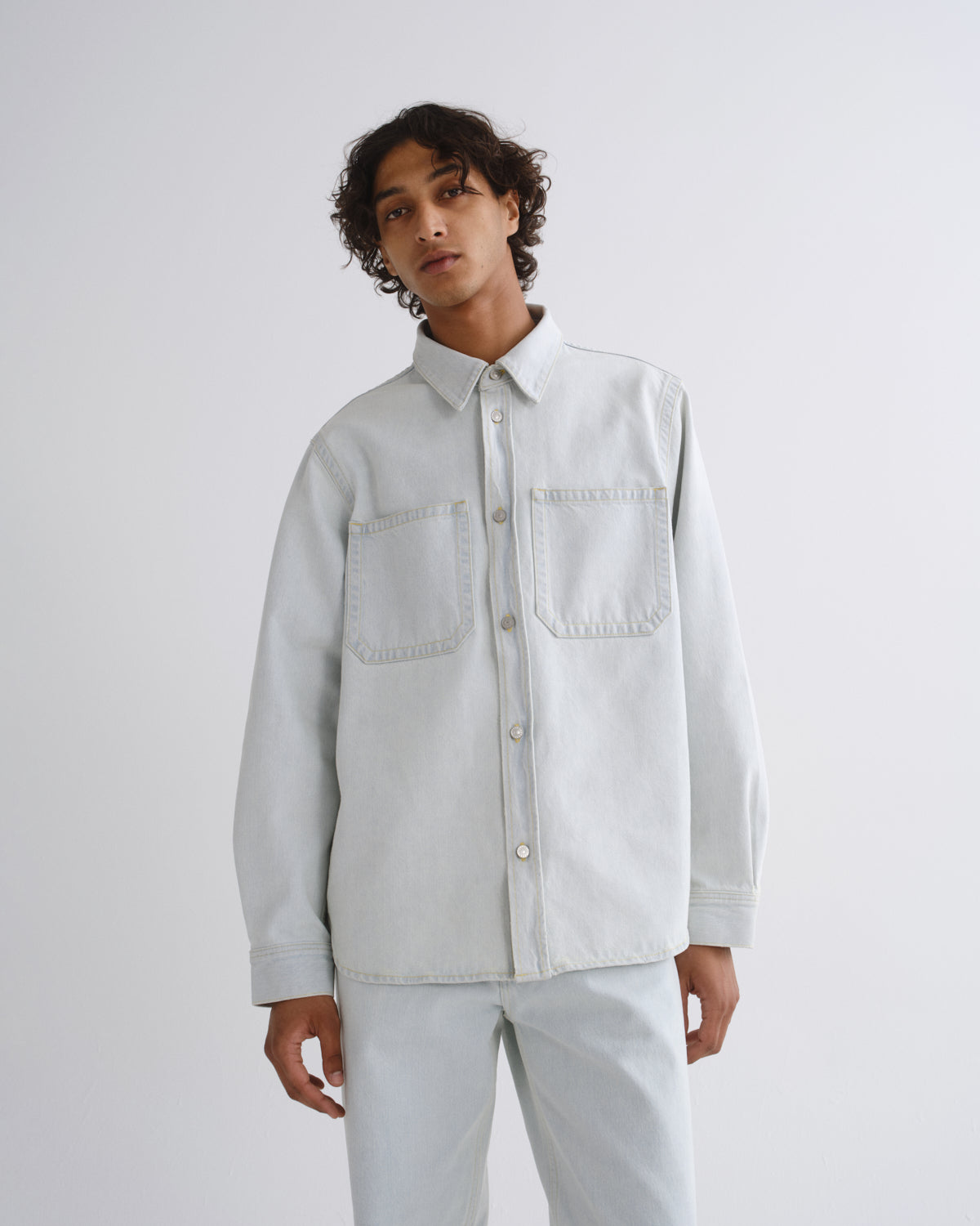 A model wearing a light gray denim button-up shirt with two chest pockets and matching light gray jeans, standing against a plain light gray studio background.