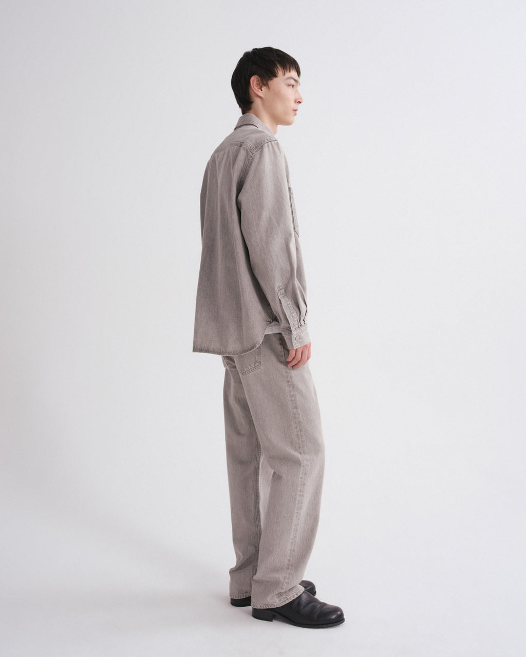 A model shown from a side profile wearing a light taupe-gray corduroy shirt and matching wide-leg trousers, paired with black loafers, against a plain white studio background.