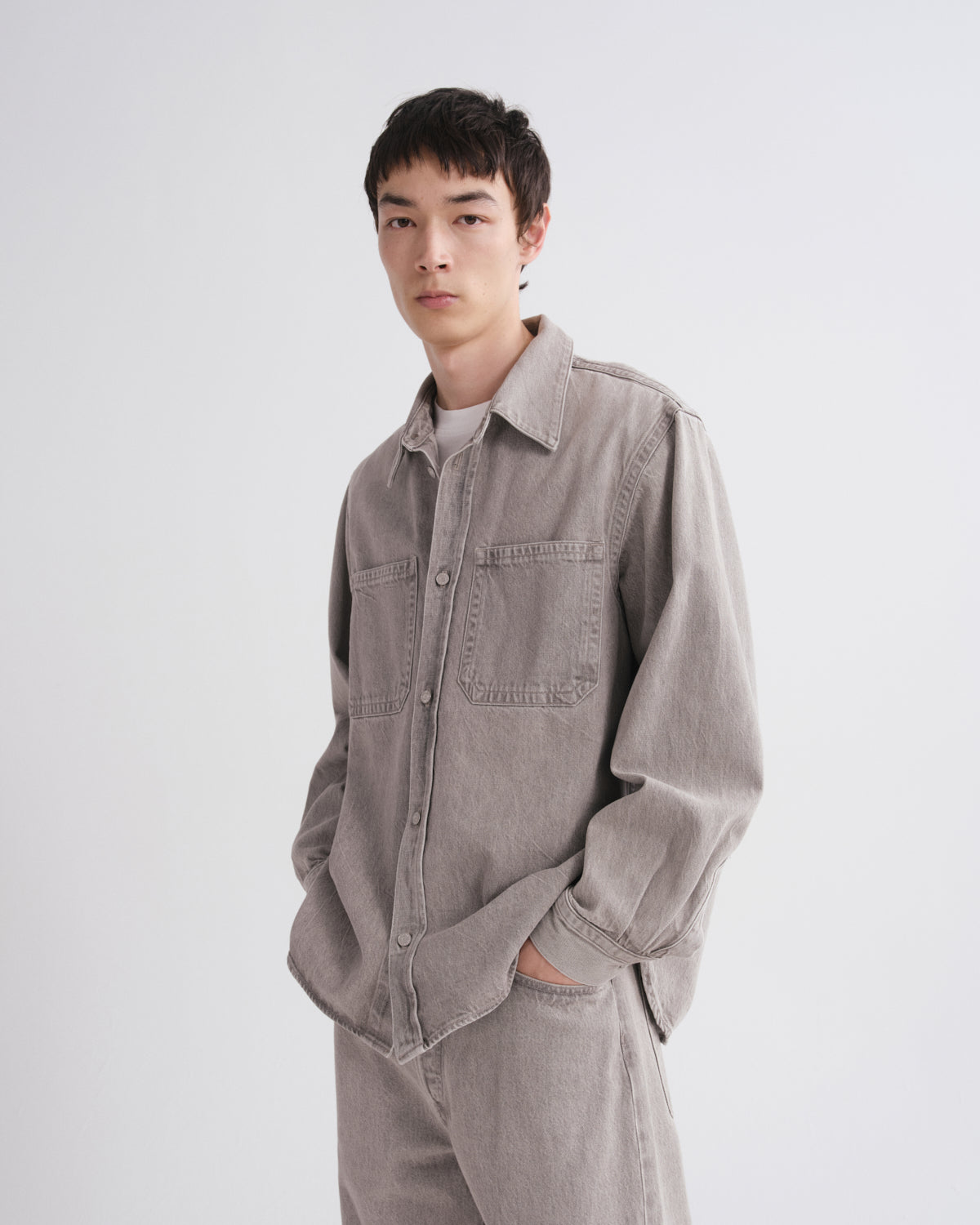 A model wearing a light gray oversized button-up shirt and matching gray pants, standing against a plain light gray studio background.