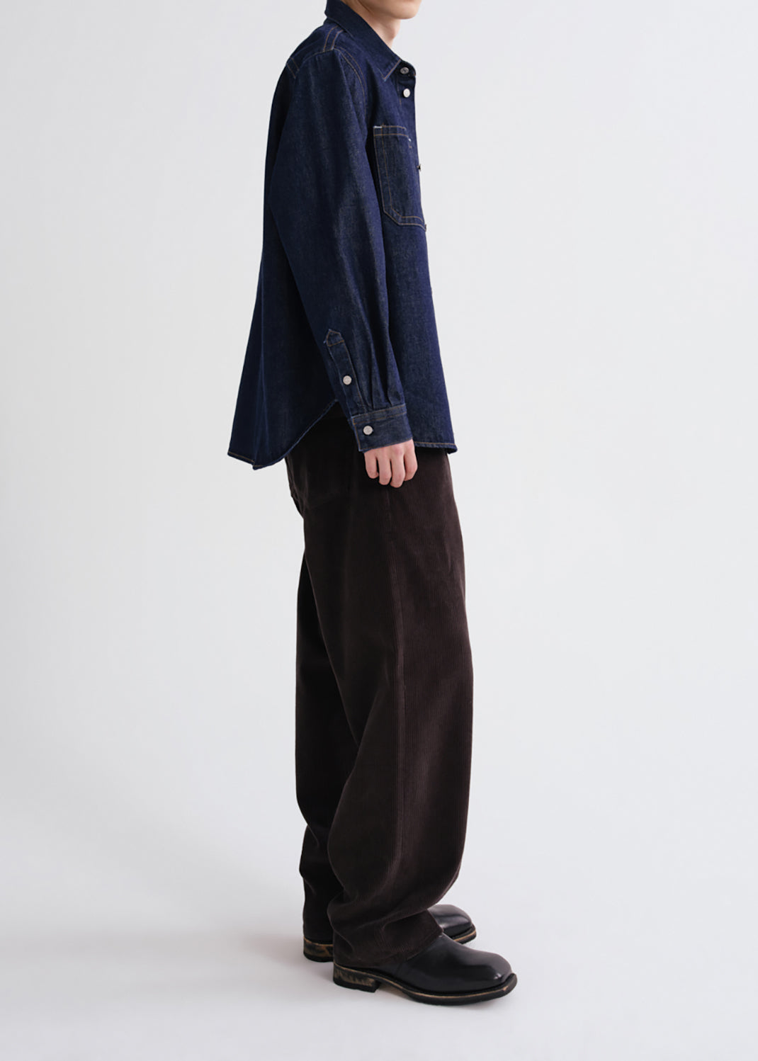A model shown from the side wearing a dark indigo denim shirt, loose dark brown corduroy trousers, and black leather boots against a grey background.